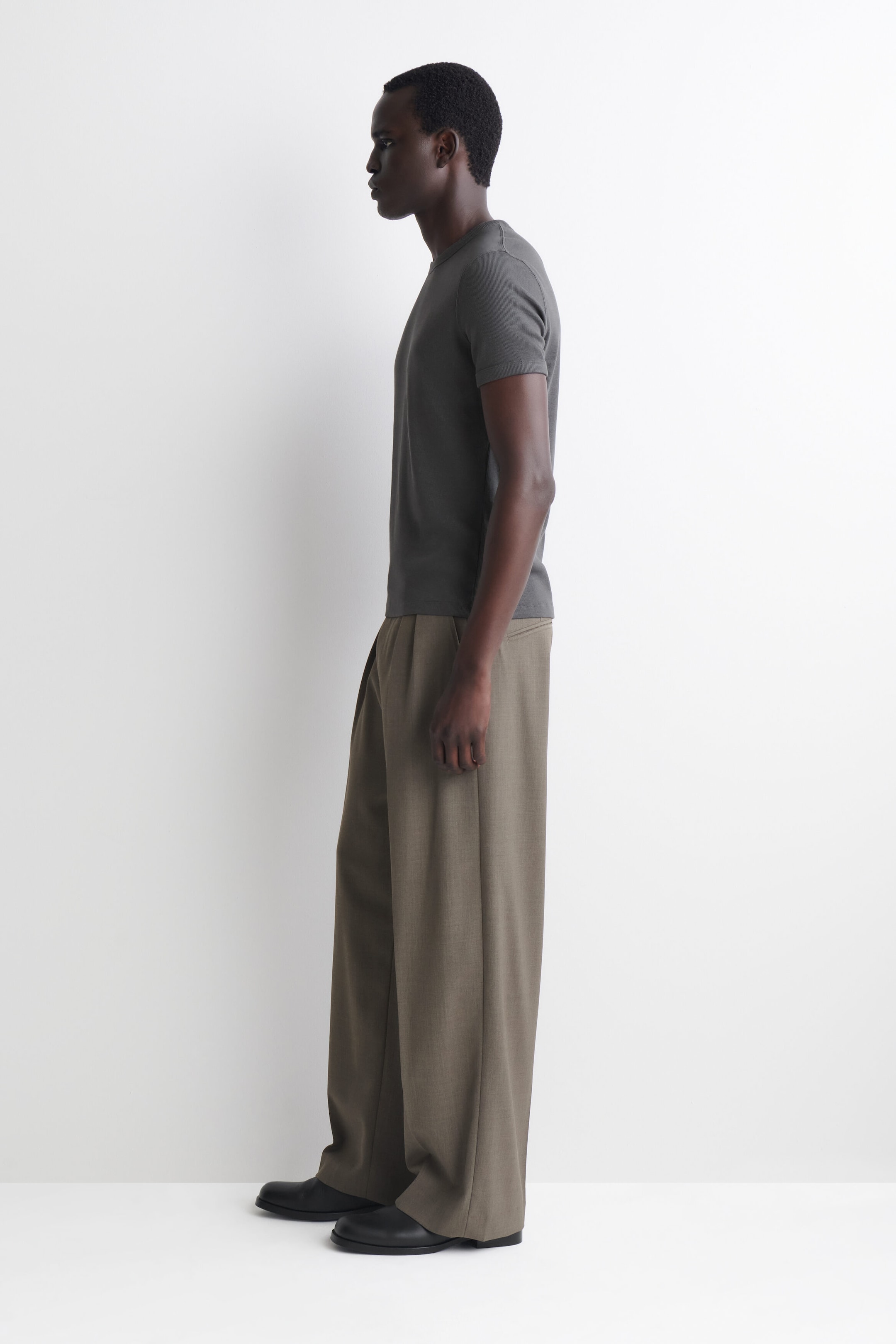 A slim ribbed cotton T-shirt in dark grey is shown in profile, facing left, highlighting its close fit, crew neck and short sleeves. The T-shirt is worn untucked over wide-leg trousers, emphasising its clean, straight hemline.