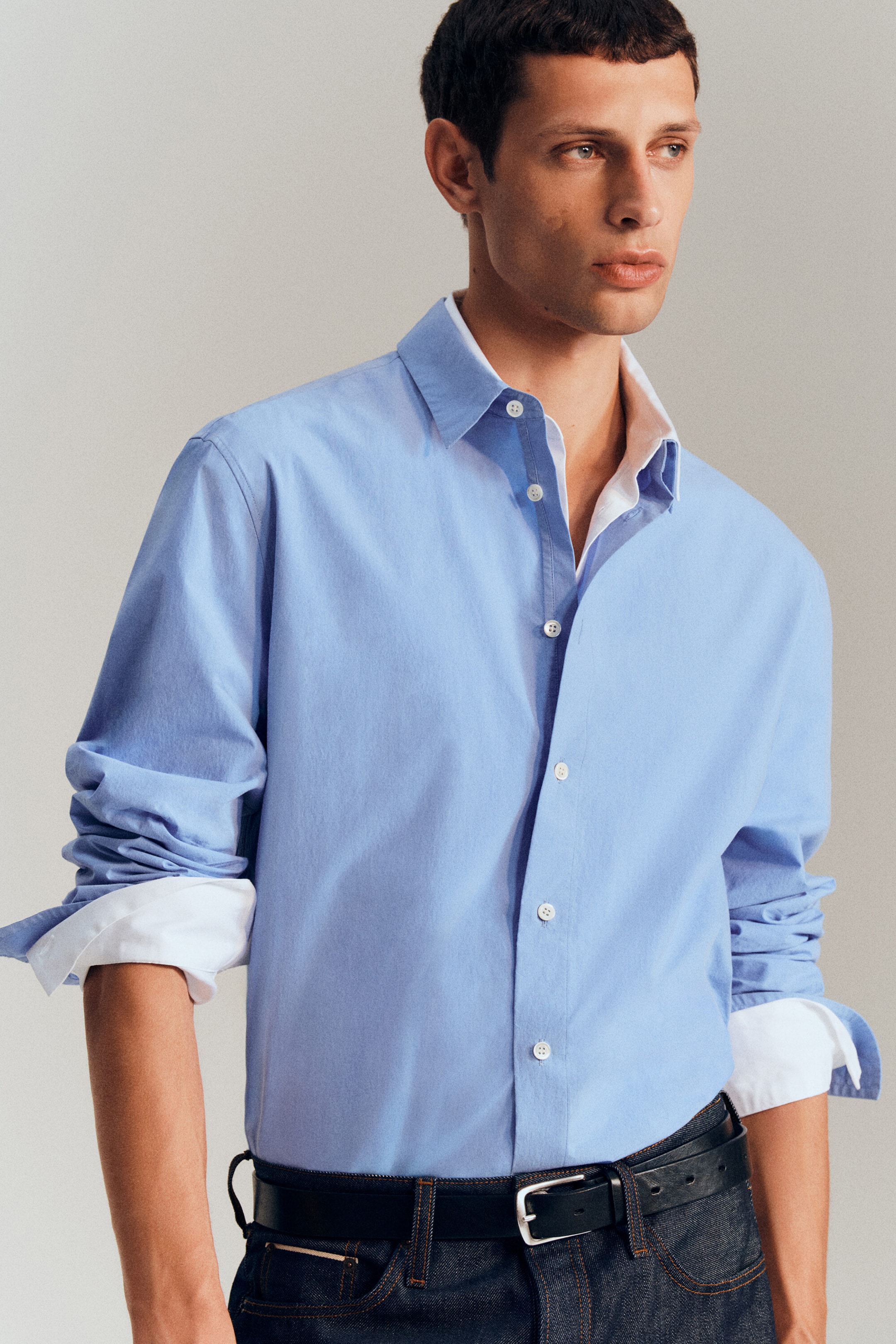 A light blue long-sleeved shirt is shown from the front, featuring a relaxed fit and a button fastening with white buttons. The shirt’s collar and rolled-up sleeves reveal a white contrast lining. A light blue long-sleeved shirt is shown from the front, featuring a relaxed fit and a button fastening with white buttons. The shirt’s collar and rolled-up sleeves reveal a white contrast lining.