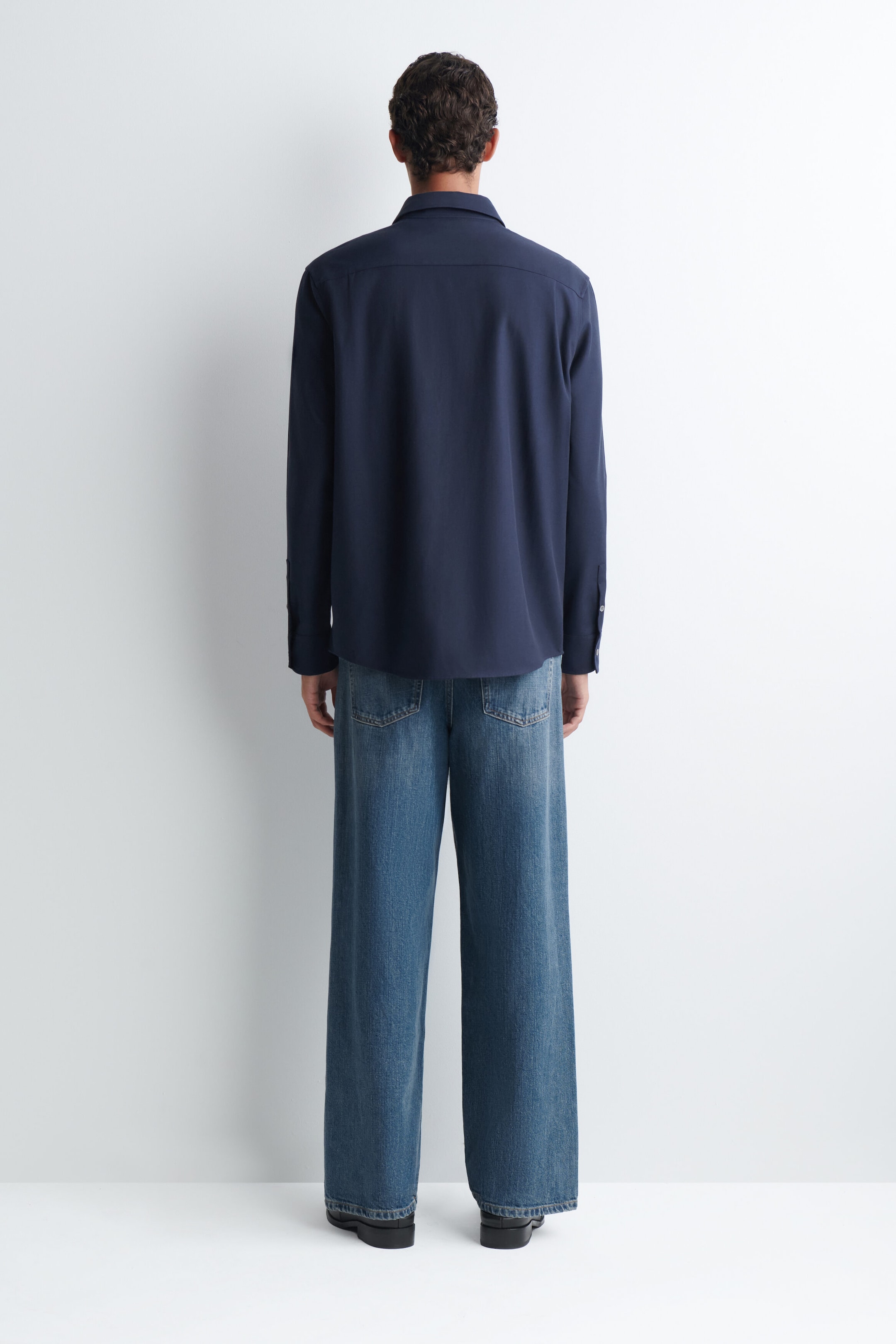 A relaxed twill shirt in navy blue is being worn by the model, shown from the back. The long-sleeved shirt features a straight hem and a collar, and is paired with wide-leg blue jeans.