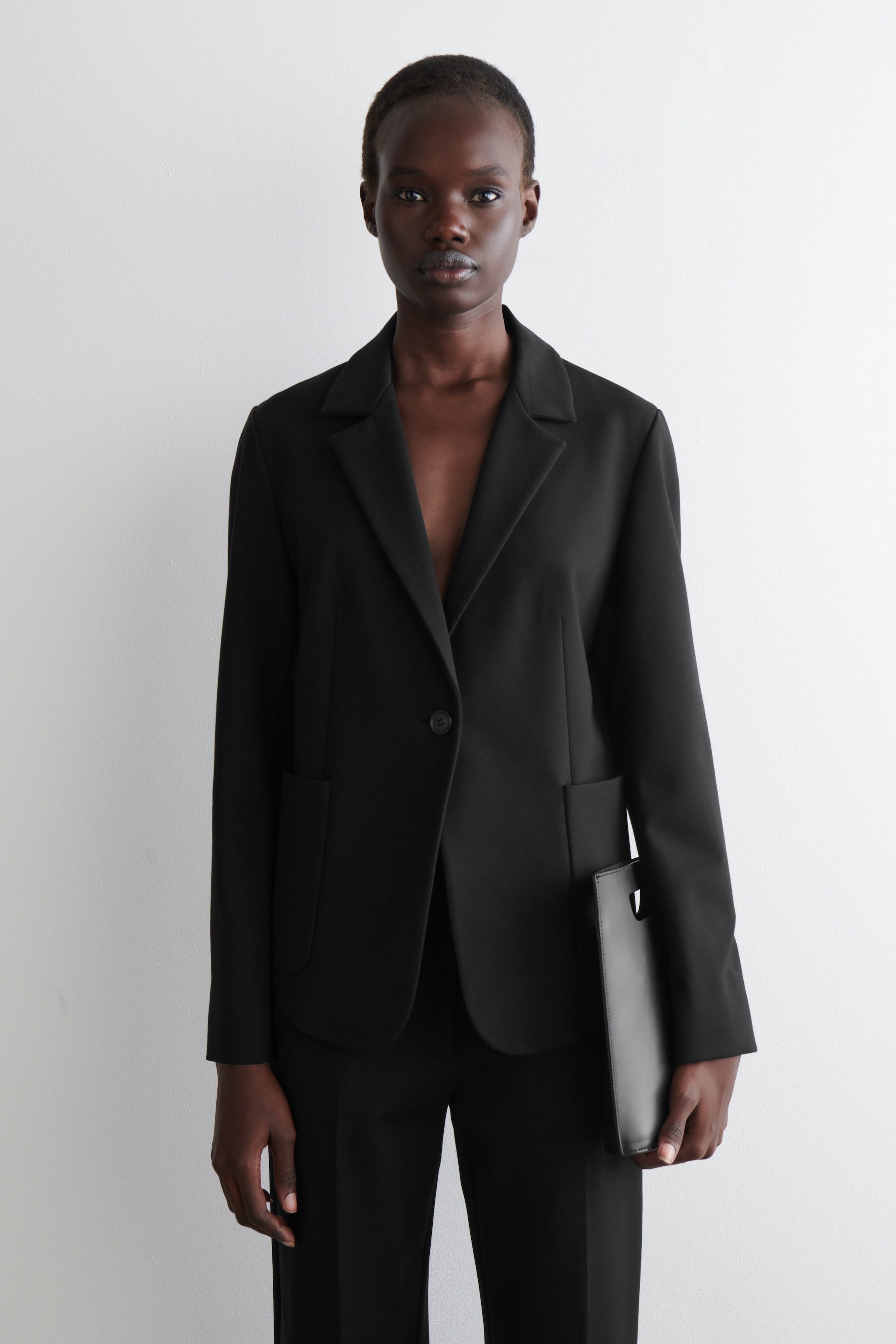 Agrandir l'image: Un blazer noir est présenté de face, avec des revers crantés classiques, une fermeture à un bouton et deux grandes poches plaquées à l'avant. Le blazer présente une coupe décontractée et structurée avec des manches longues.