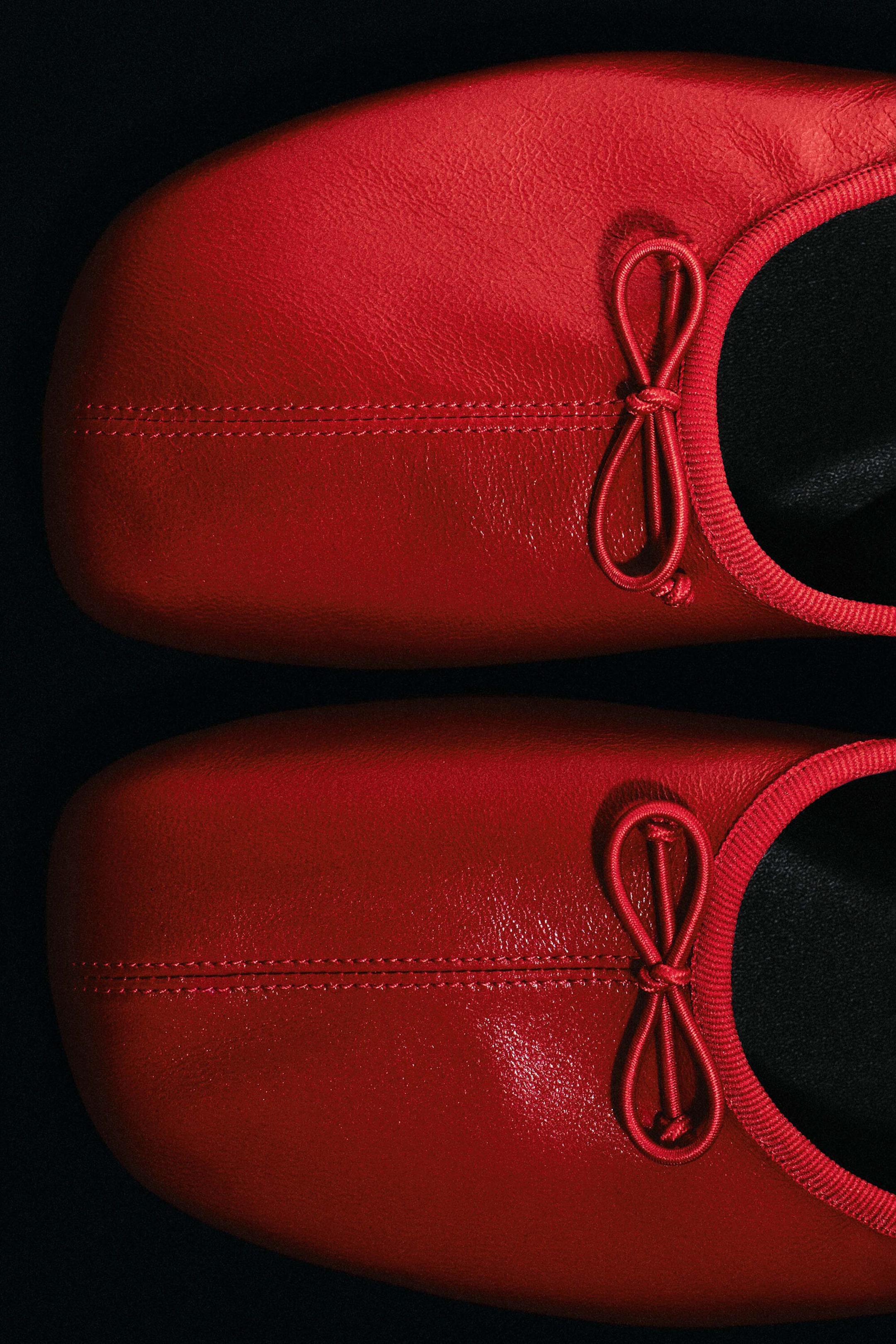 A pair of red leather ballet flats is shown from a close-up overhead angle, highlighting their square-toe design. The flats feature a central top-stitched seam on each toe, matching red grosgrain trim along the collar, and small decorative bows.