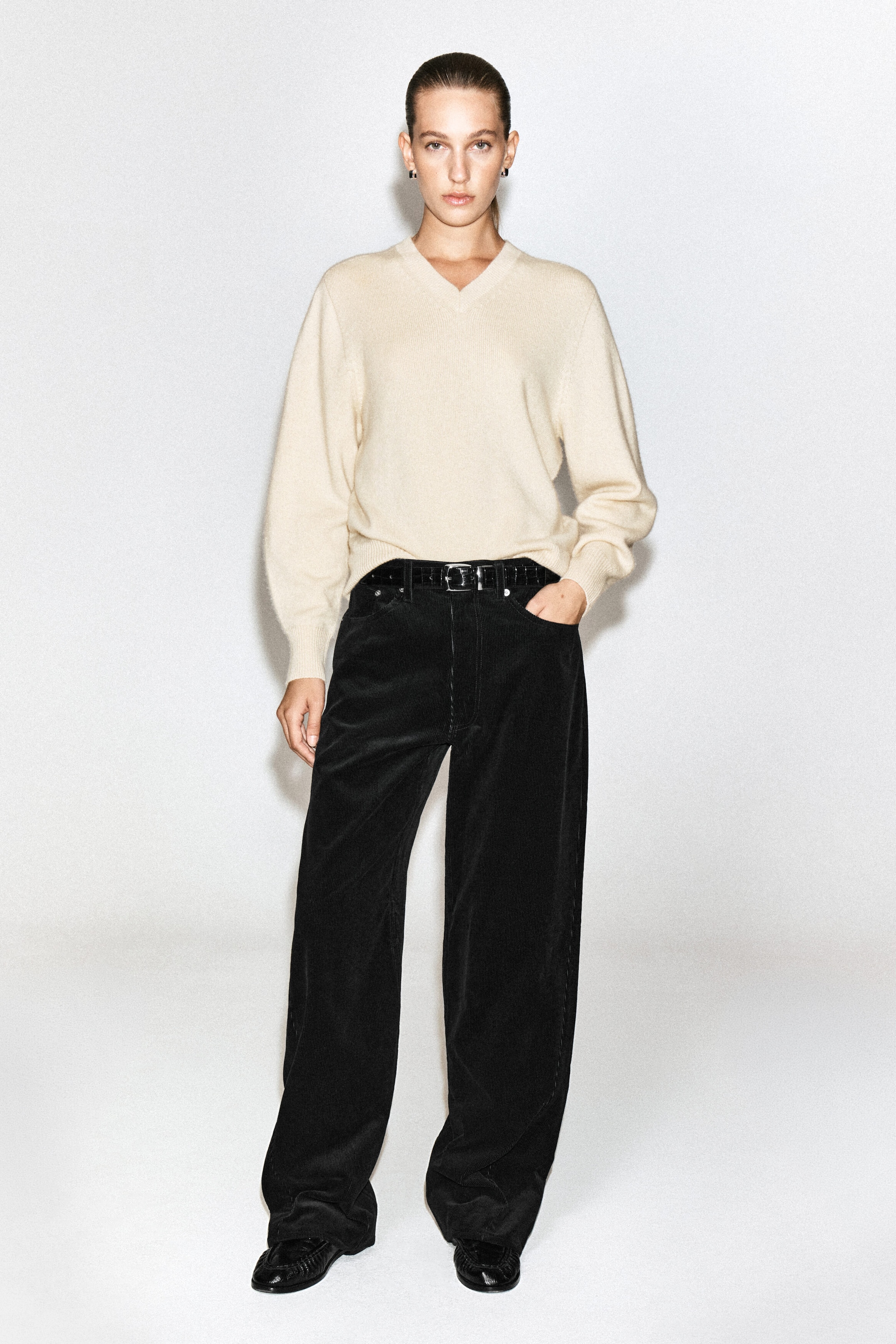 The model is wearing these black corduroy straight-leg jeans, standing with one hand resting in the front pocket. The high-waisted jeans are styled with a black belt and a tucked-in cream-coloured V-neck sweater. The model is wearing these black corduroy straight-leg jeans, standing with one hand resting in the front pocket. The high-waisted jeans are styled with a black belt and a tucked-in cream-coloured V-neck sweater.