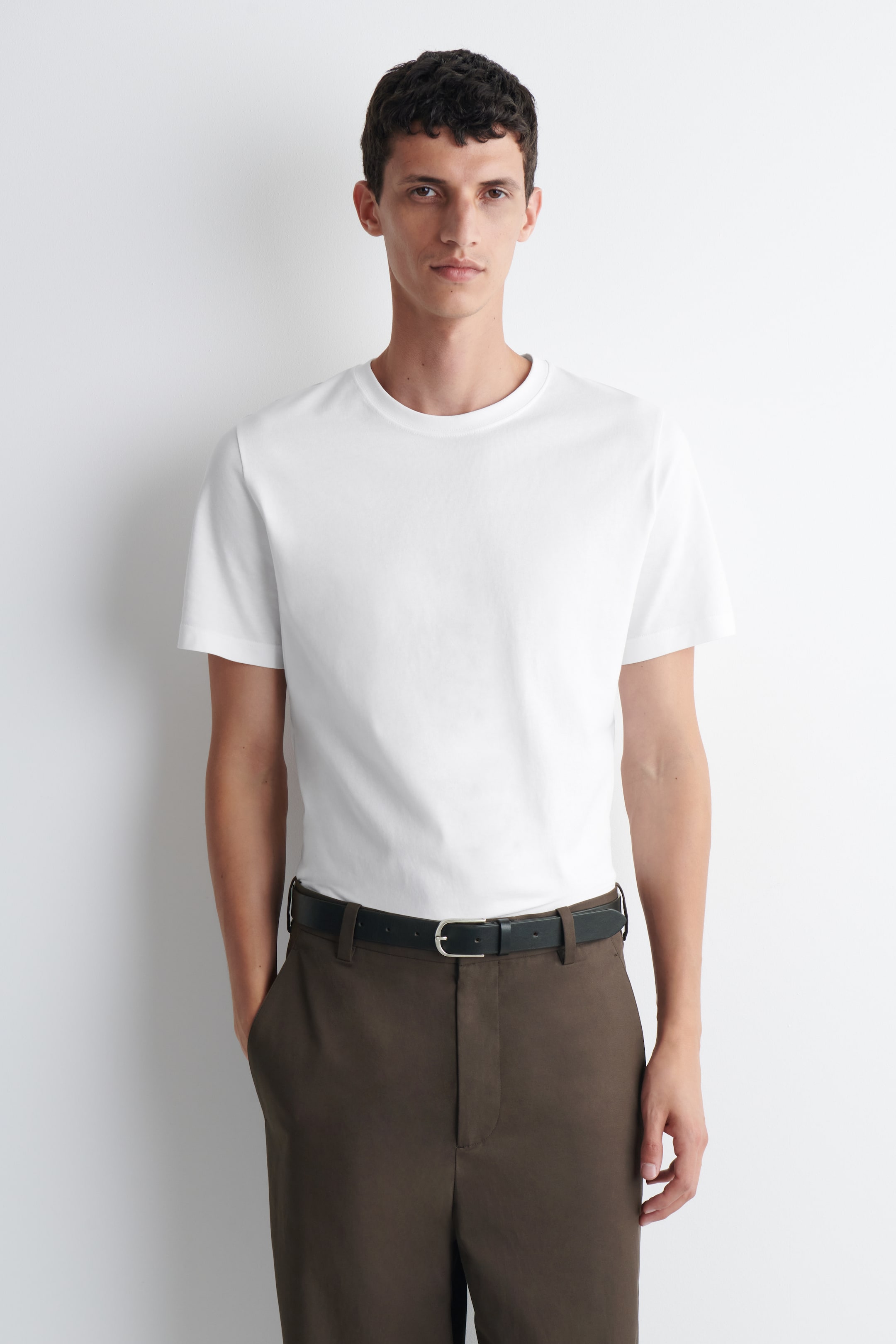 Grotere afbeelding bekijken: Een wit T-shirt met ronde hals en korte mouwen wordt gedragen door het model. Het T-shirt is ingestopt in een bruine broek.