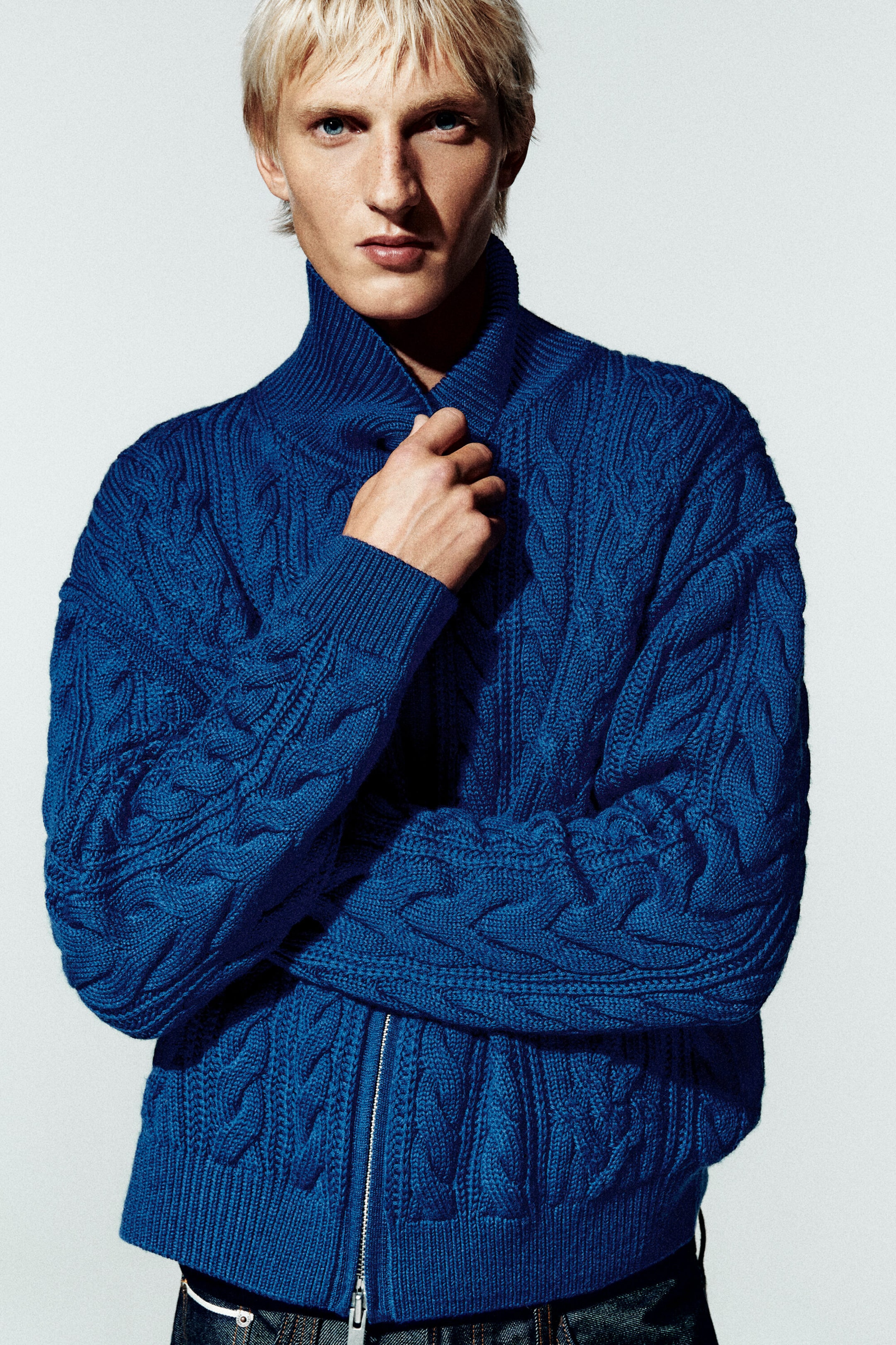 A royal blue cable-knit jacket is shown from the front, featuring a textured cable pattern, a high funnel neck collar, and a silver two-way zipper. The model is wearing the regular-fit jacket zipped up, highlighting its ribbed cuffs and hem. A royal blue cable-knit jacket is shown from the front, featuring a textured cable pattern, a high funnel neck collar, and a silver two-way zipper. The model is wearing the regular-fit jacket zipped up, highlighting its ribbed cuffs and hem.