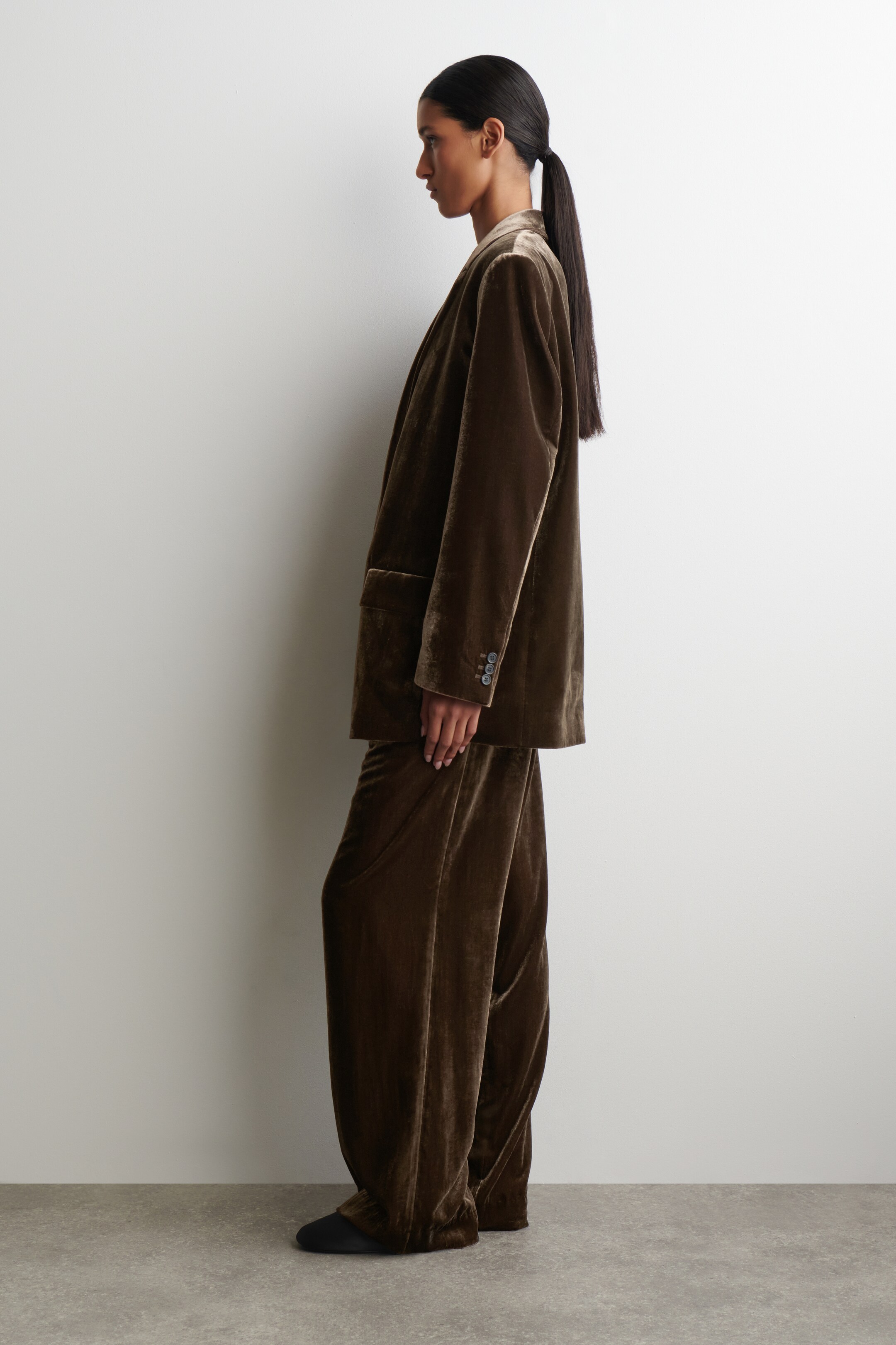 Agrandir l'image: Un costume en velours brun olive, comprenant un blazer et un pantalon large, est présenté face à gauche. Le blazer est détaillé avec des revers crantés, une poche plaquée sur la hanche et trois boutons sur sa manche longue, tandis que le pantalon est conçu avec une coupe ample et longue.