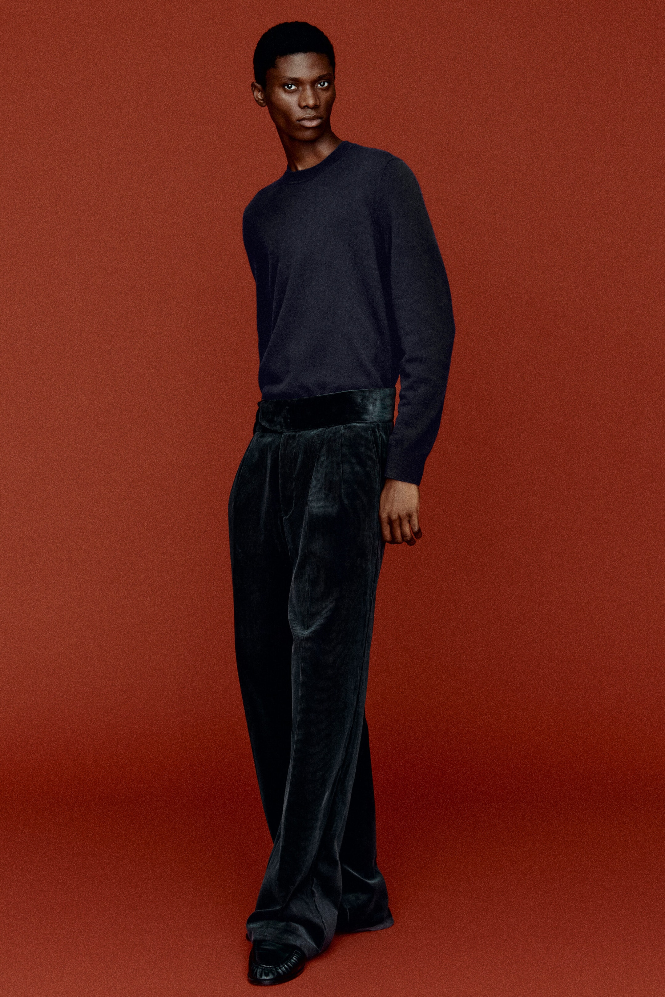 Un chandail en cachemire bleu marine à col ras du cou est porté rentré dans un pantalon en velours côtelé foncé à jambes larges, présentant la tenue sous un angle frontal à trois quarts. Le pantalon présente une taille haute inspirée du cummerbund et des plis devant qui tombent dans un drapé décontracté.