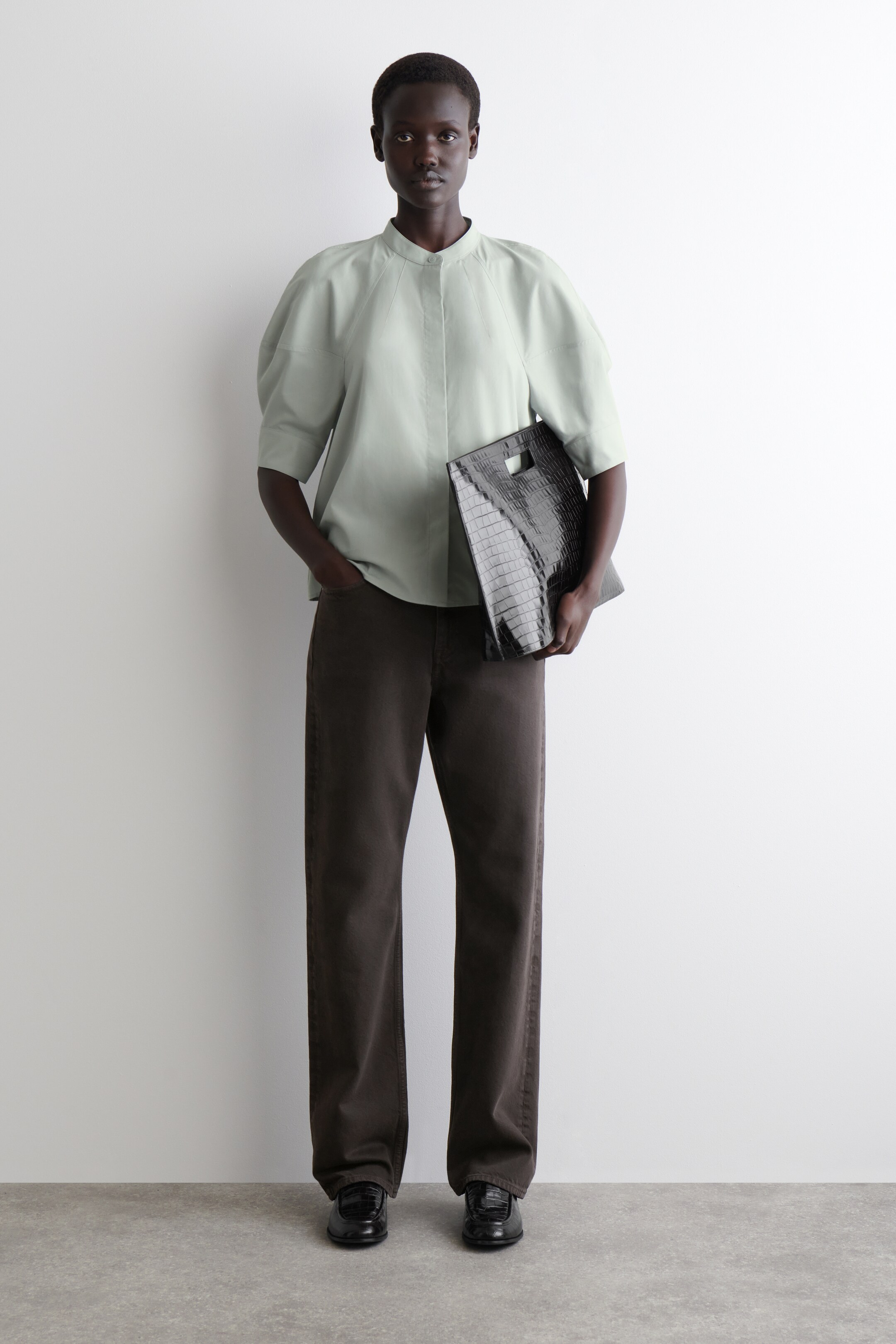 Grotere afbeelding bekijken: Een lichtgroene blouse met knopen aan de voorkant, voorzien van een bandkraag en volumineuze raglanmouwen tot aan de elleboog, wordt gedragen met donkerbruine wijde broek. Een zwarte clutch tas met krokodillenprint en een uitsnede handvat wordt vastgehouden, en zwarte glanzende loafers maken de look compleet.
