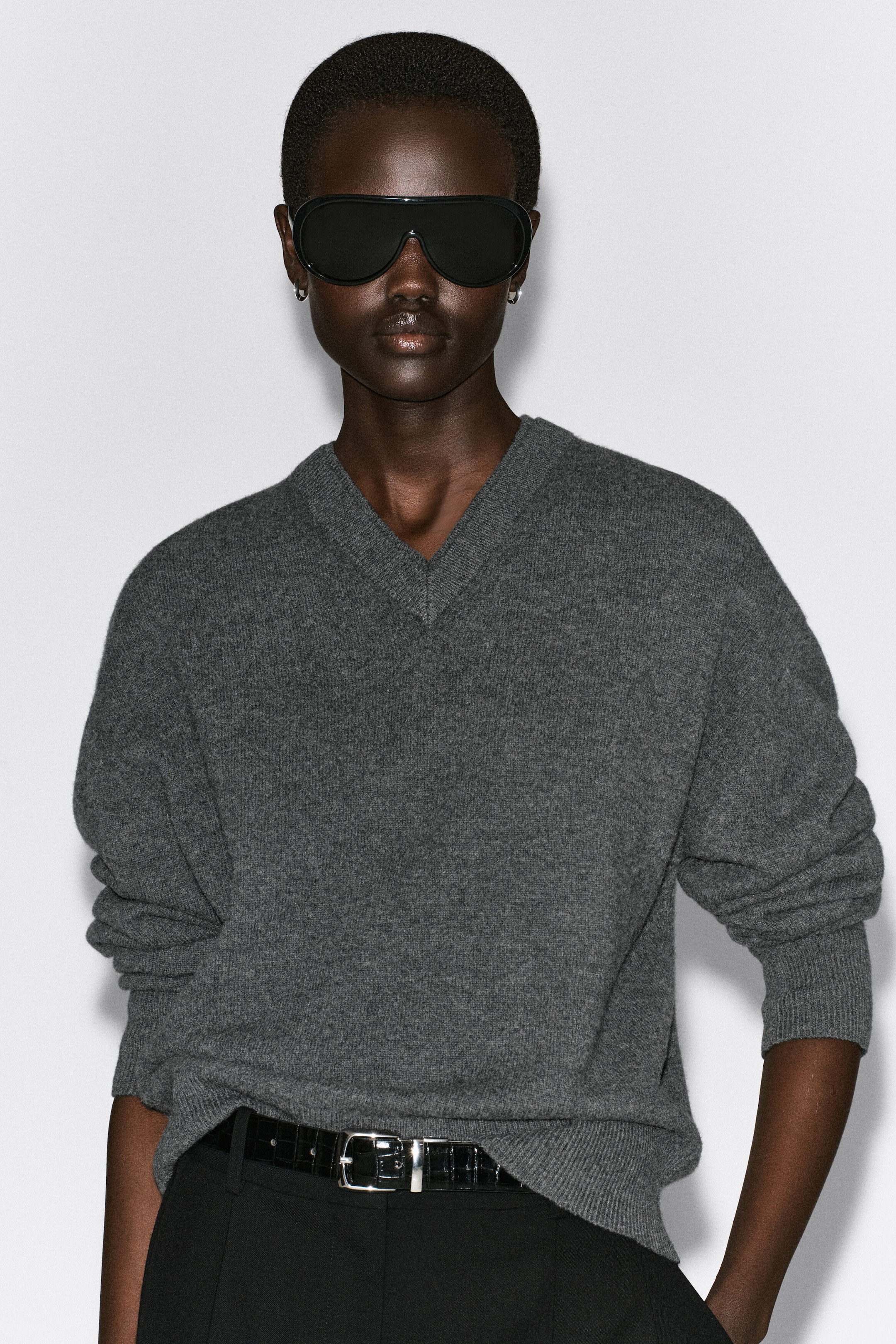 This front view of the gray merino wool sweater showcases a relaxed fit with dropped shoulders, a layered ribbed V-neckline, and long sleeves gathered at the ribbed cuffs. The top drapes softly over the torso, with the hem partially tucked into a belted waistband to define its slouchy silhouette. This front view of the gray merino wool sweater showcases a relaxed fit with dropped shoulders, a layered ribbed V-neckline, and long sleeves gathered at the ribbed cuffs. The top drapes softly over the torso, with the hem partially tucked into a belted waistband to define its slouchy silhouette.