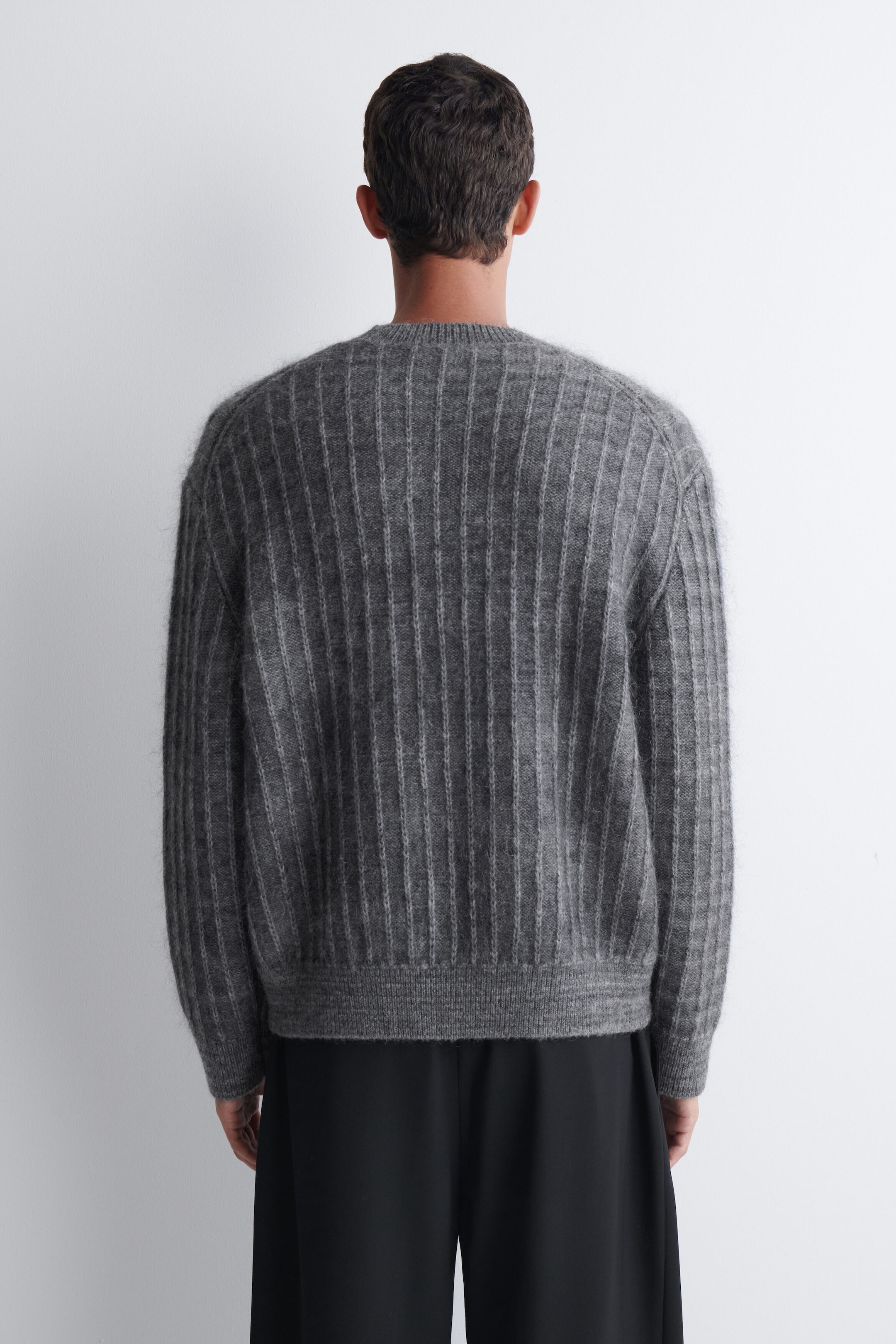 A grey rib-knit sweater is being worn by the model, viewed from the back. The sweater features a fuzzy texture with vertical ribbing, ribbed trims at the hem and cuffs, and is styled with black trousers. A grey rib-knit sweater is being worn by the model, viewed from the back. The sweater features a fuzzy texture with vertical ribbing, ribbed trims at the hem and cuffs, and is styled with black trousers.