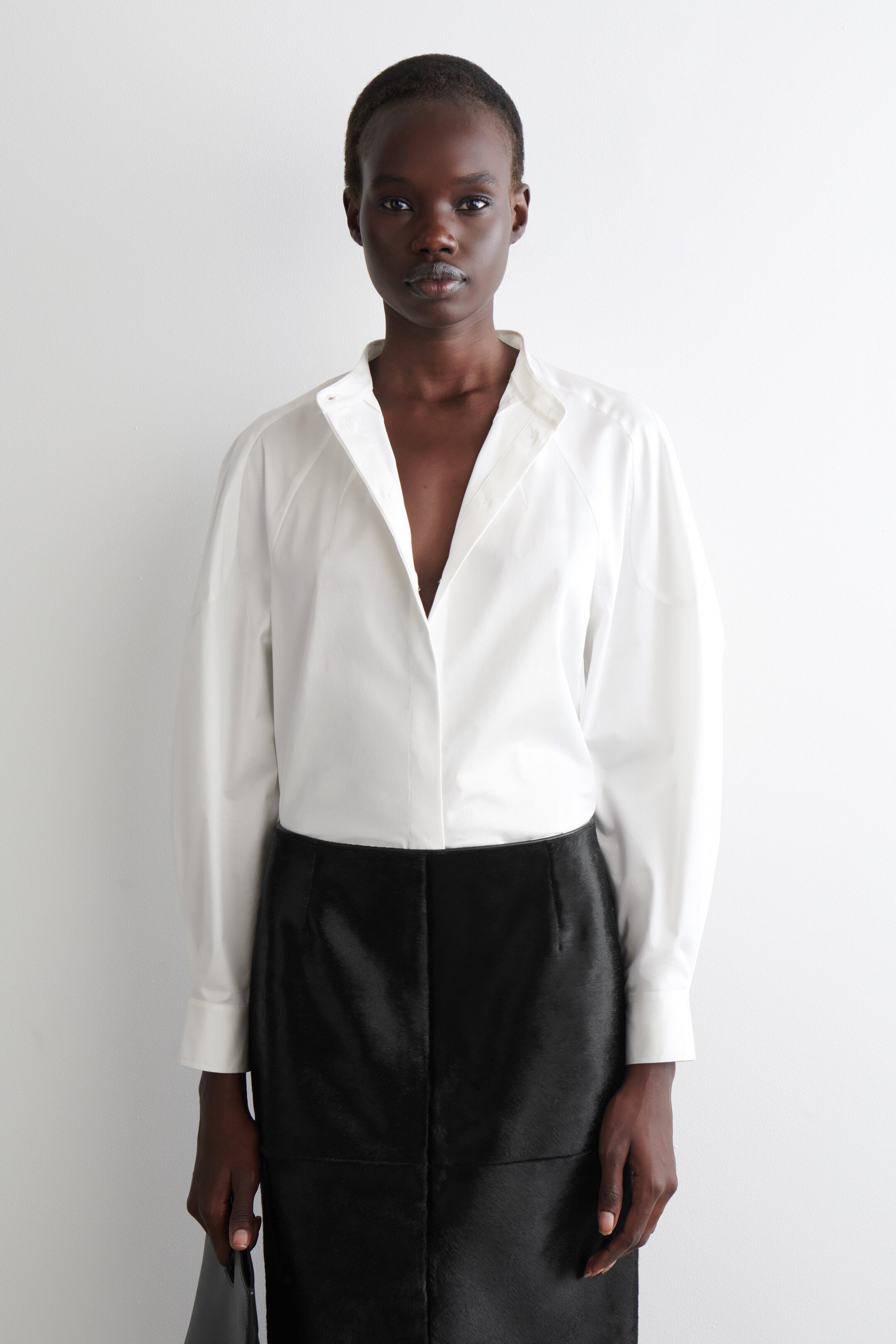 View larger image: A white, relaxed-fit shirt is being shown front-facing, featuring a stand collar that is open to form a deep V-neck and long raglan sleeves with button cuffs. The shirt is being tucked into a black, high-waisted skirt with visible paneling and a subtle texture.