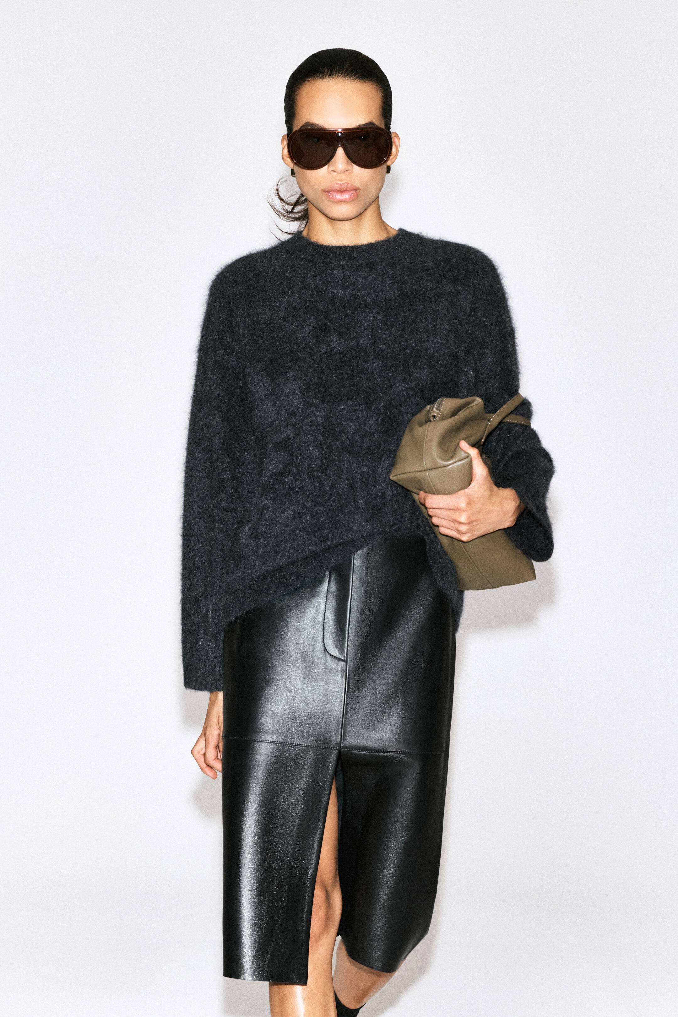 Größeres Bild ansehen: Das Outfit besteht aus einem dunkelgrauen, flauschigen Oversize-Pullover mit langen Ärmeln, kombiniert mit einem schwarzen, knielangen Lederrock. Der Rock zeigt einen geraden Schnitt mit einem markanten Schlitz vorne.