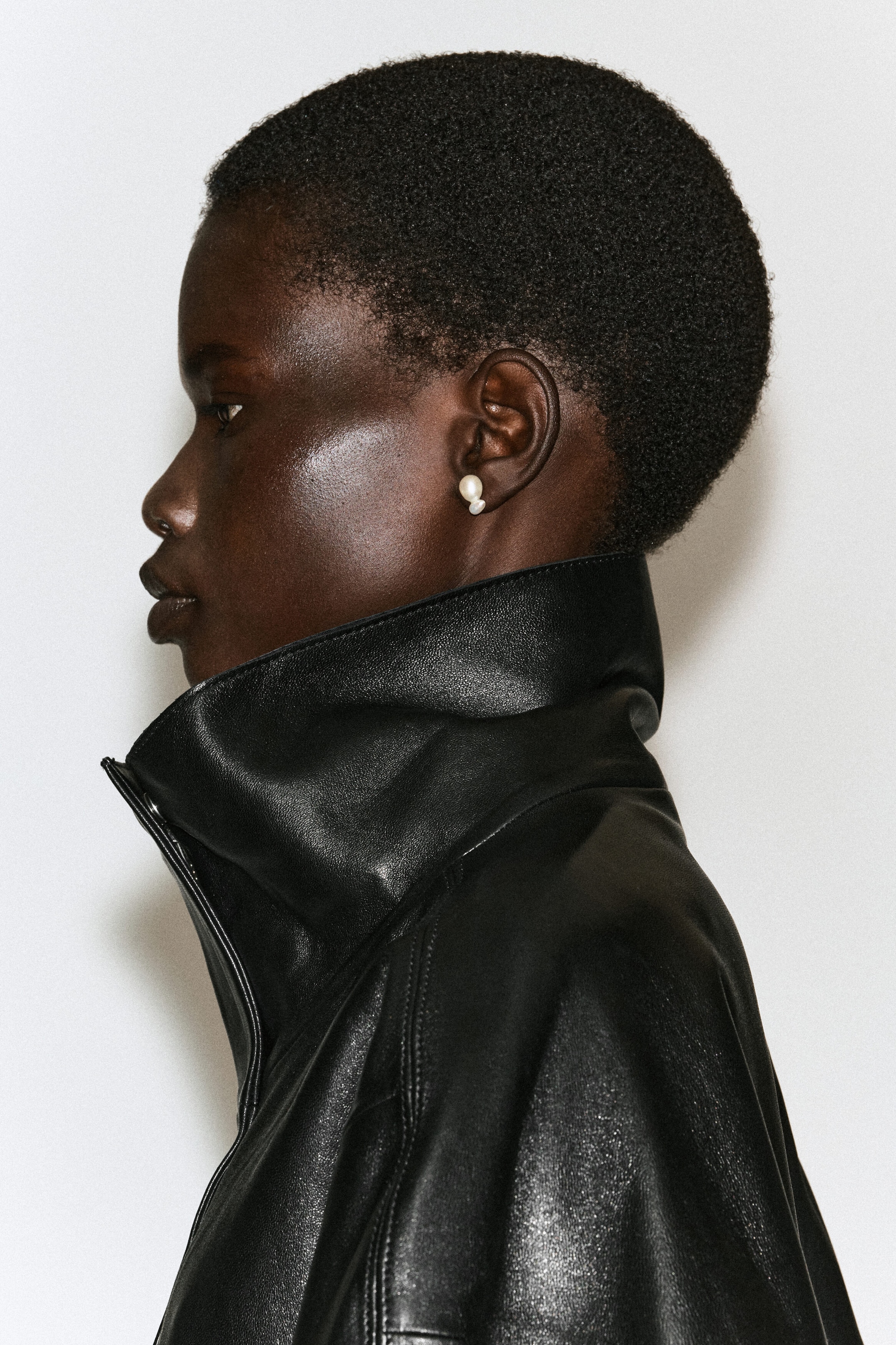A sculptural freshwater pearl earring, featuring an organic double-lobed white shape, is worn on the model's earlobe. The earring is styled with a black leather jacket featuring a high, structured collar. A sculptural freshwater pearl earring, featuring an organic double-lobed white shape, is worn on the model's earlobe. The earring is styled with a black leather jacket featuring a high, structured collar.