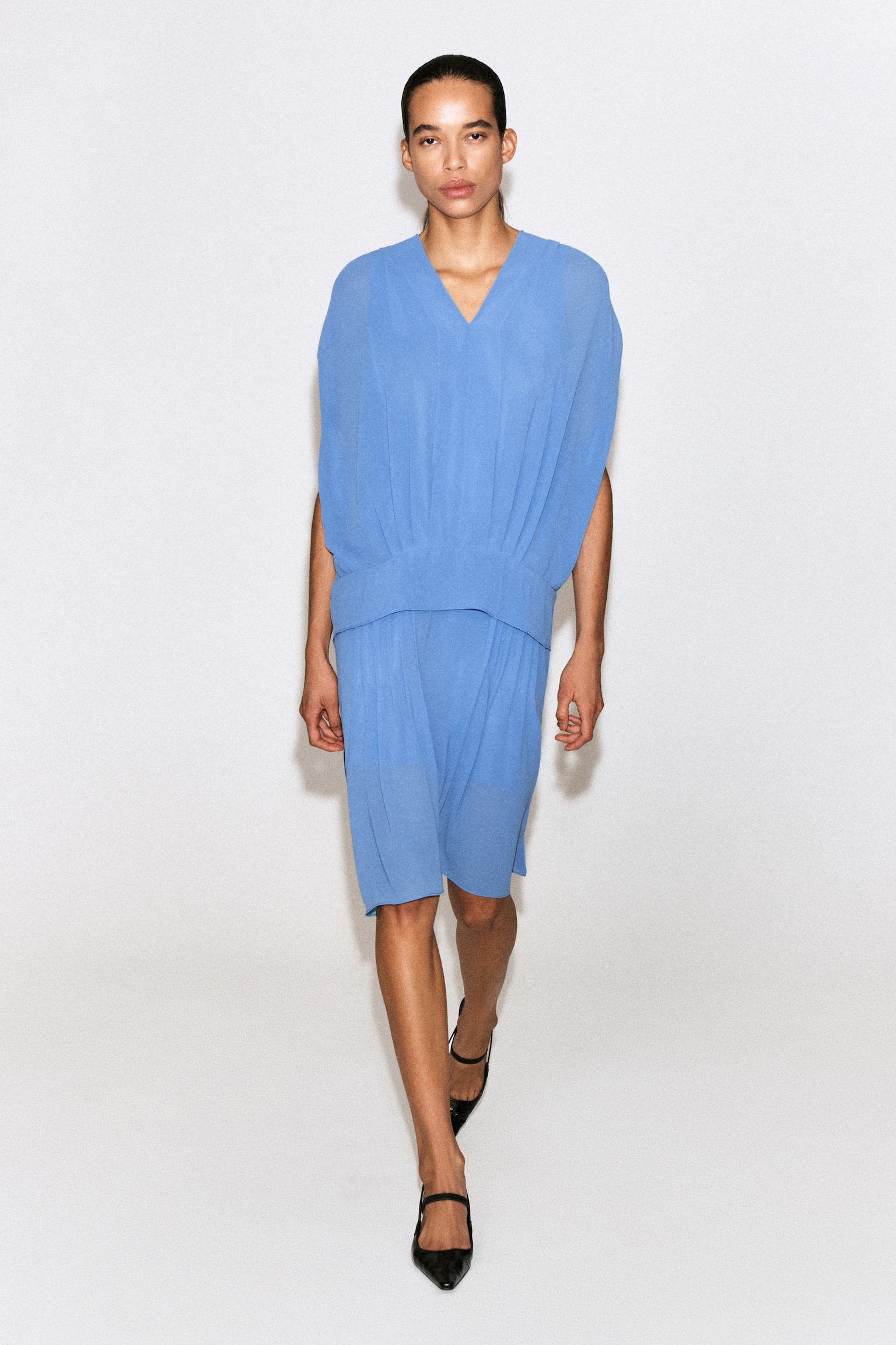 Ein hellblaues, drapiertes Midi-Kleid mit Falten und V-Ausschnitt wird von vorne gezeigt. Es verfügt über einen V-Ausschnitt und breite, drapierte Schultern, die eine ärmellose Silhouette erzeugen. Das Kleid ist mit einem breiten, taillierten Bund und einem plissierten, knielangen Rock aus halbtransparentem Stoff gestaltet. - 1
