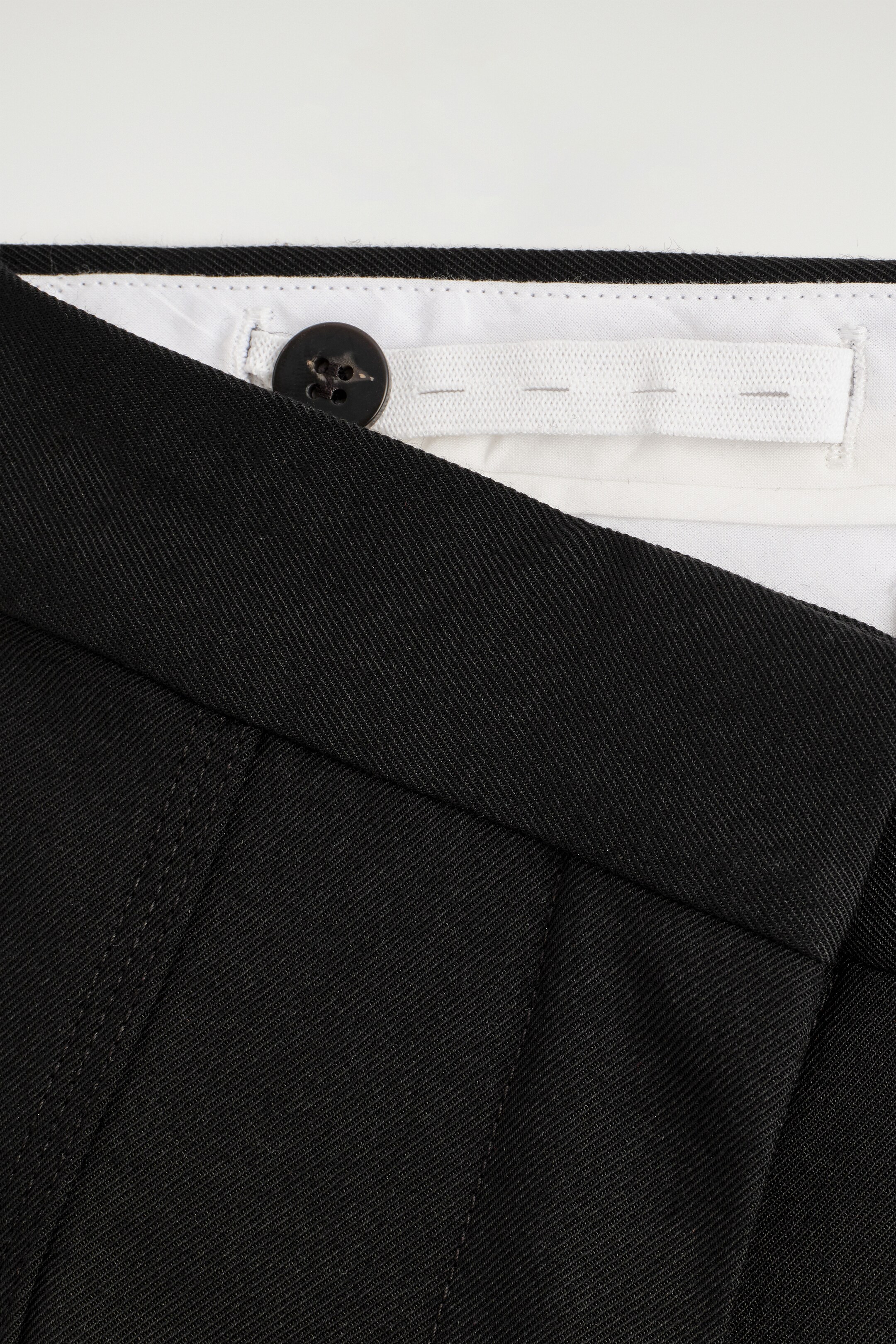 Vezi imaginea mai mare: Pantalonii din țesătură twill neagră sunt prezentați din față, având un nasture închis la culoare care închide talia și pliuri frontale proeminente. O bandă elastică albă cu mai multe butoniere este vizibilă în interiorul taliei, indicând o potrivire ajustabilă.