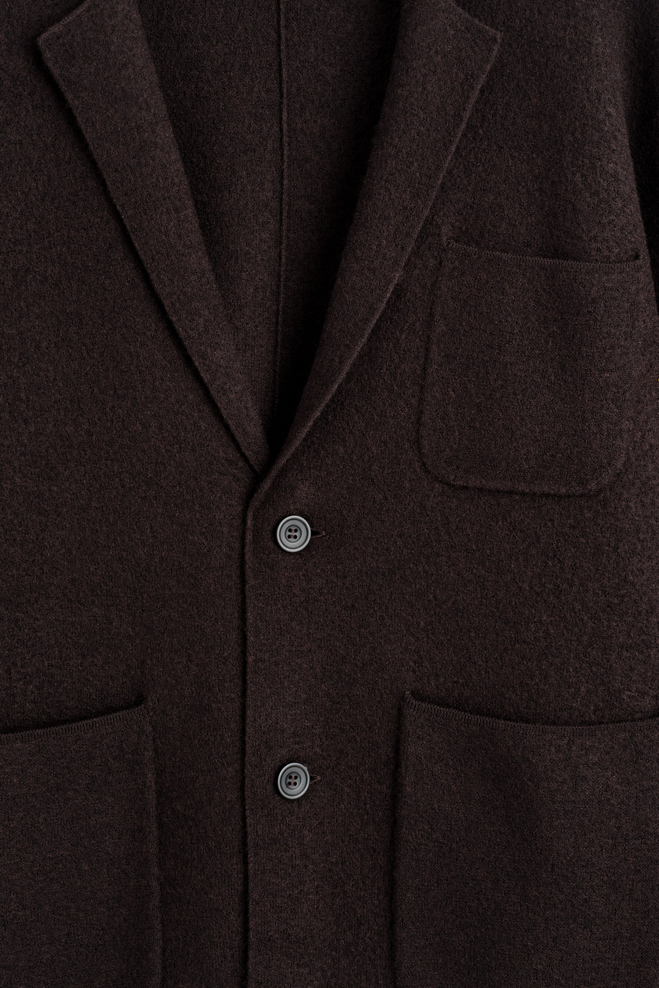 Ver imagem maior: Este blazer castanho escuro está a ser apresentado de frente, apresentando um tecido texturizado de lã merino fervida. É desenhado com lapelas clássicas em bico, dois botões tonais visíveis e dois grandes bolsos frontais de patch.