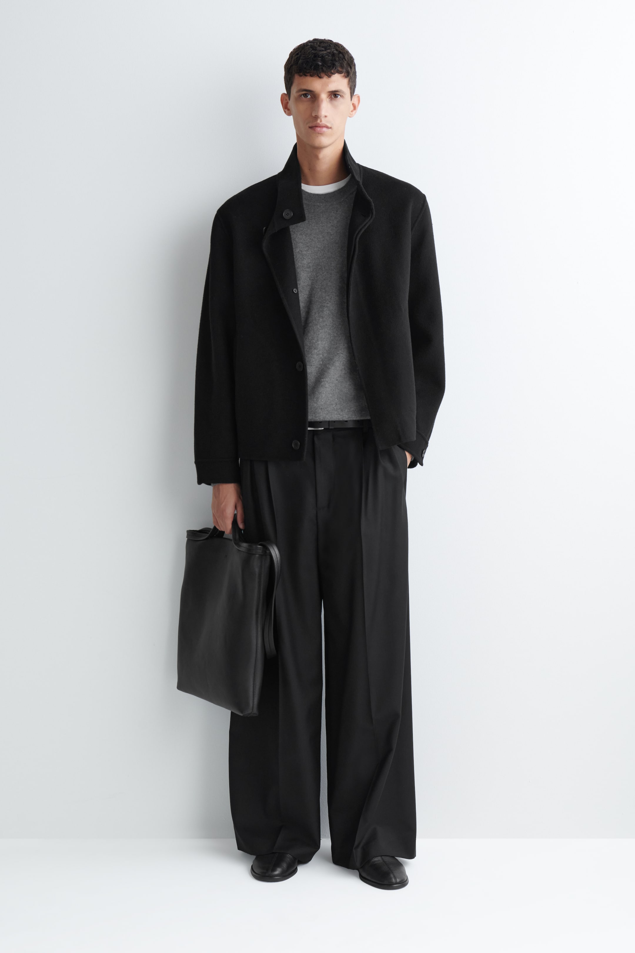 Grotere afbeelding bekijken: Plooiige wide-leg broek in relaxed fit in zwart, gedragen door het model, met plooien aan de voorkant en een losse, soepel vallende silhouet. De broek is gestyled met een smalle zwarte riem, een grijze sweater met ronde hals, een zwart jack en zwarte schoenen, terwijl een zwarte leren tas wordt gedragen.