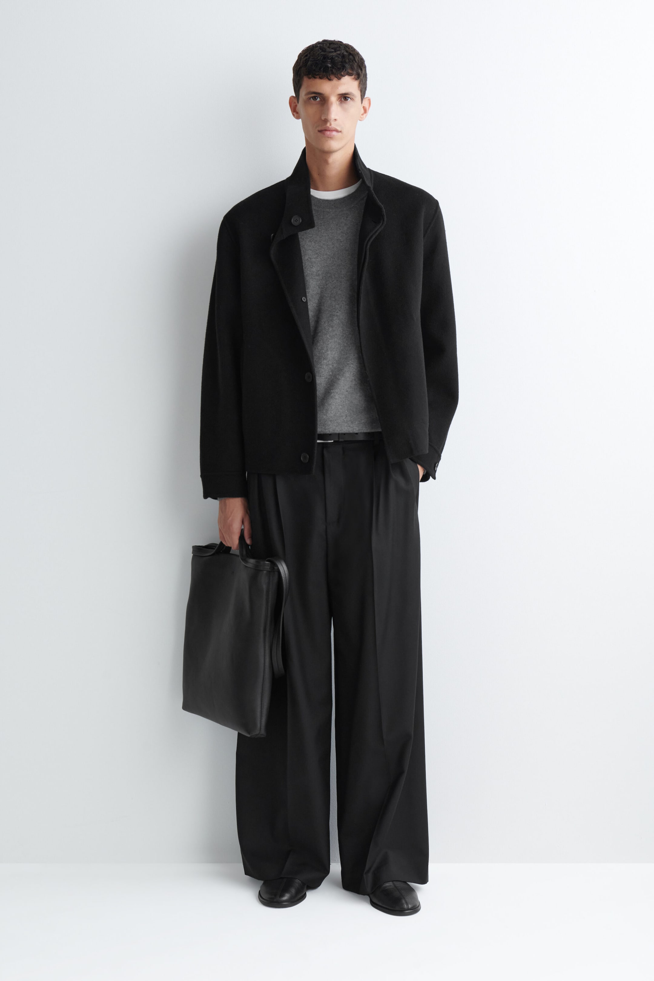 Schwarze, plissierte Wide-Leg-Hose in lockerer Passform, getragen von dem Model, mit Frontfalten und einer weiten, fließenden Silhouette. Die Hose wird mit einem schmalen schwarzen Gürtel, einem grauen Rundhalspullover, einer schwarzen Jacke und schwarzen Schuhen kombiniert; dazu wird eine schwarze Ledertasche getragen.