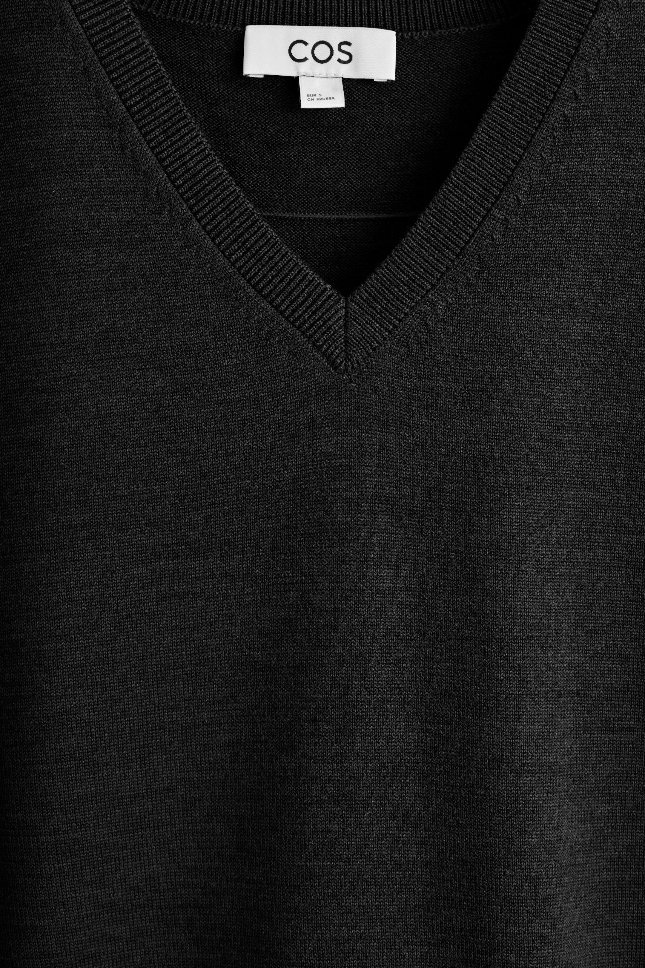 Größeres Bild ansehen: Ein schwarzer V-Ausschnitt-Pullover wird von vorne gezeigt, mit einer fein gestrickten Textur und einem gerippten V-Ausschnitt. Ein weißes Markenetikett mit 'COS' ist im Inneren des Nackens sichtbar.