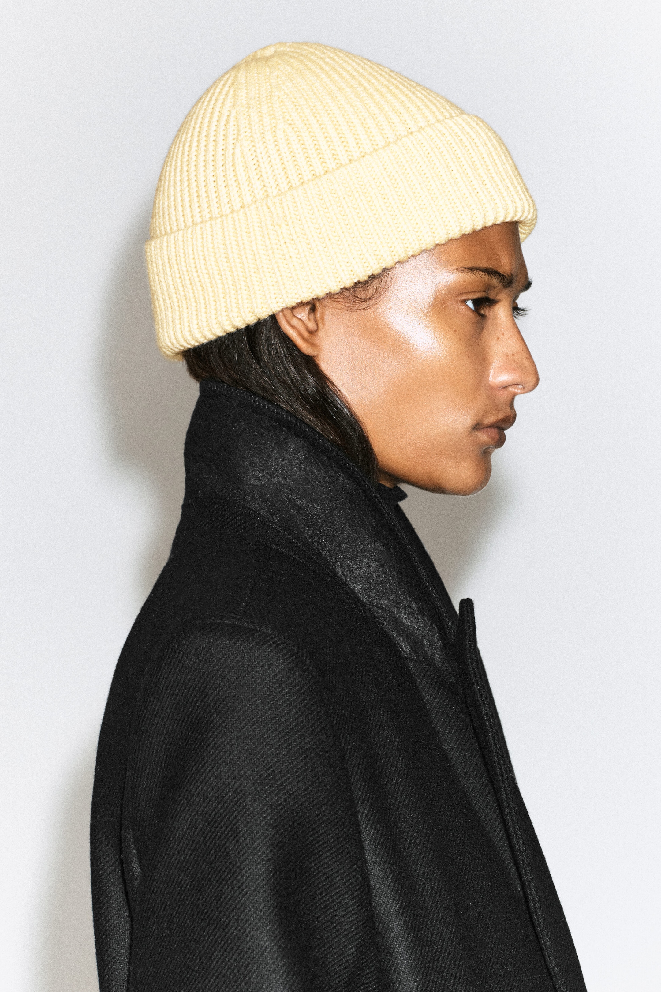 A chunky ribbed wool beanie in a light yellow shade, featuring a folded cuff, is worn on the model's head. The beanie is styled with a high-collared black coat. A chunky ribbed wool beanie in a light yellow shade, featuring a folded cuff, is worn on the model's head. The beanie is styled with a high-collared black coat.