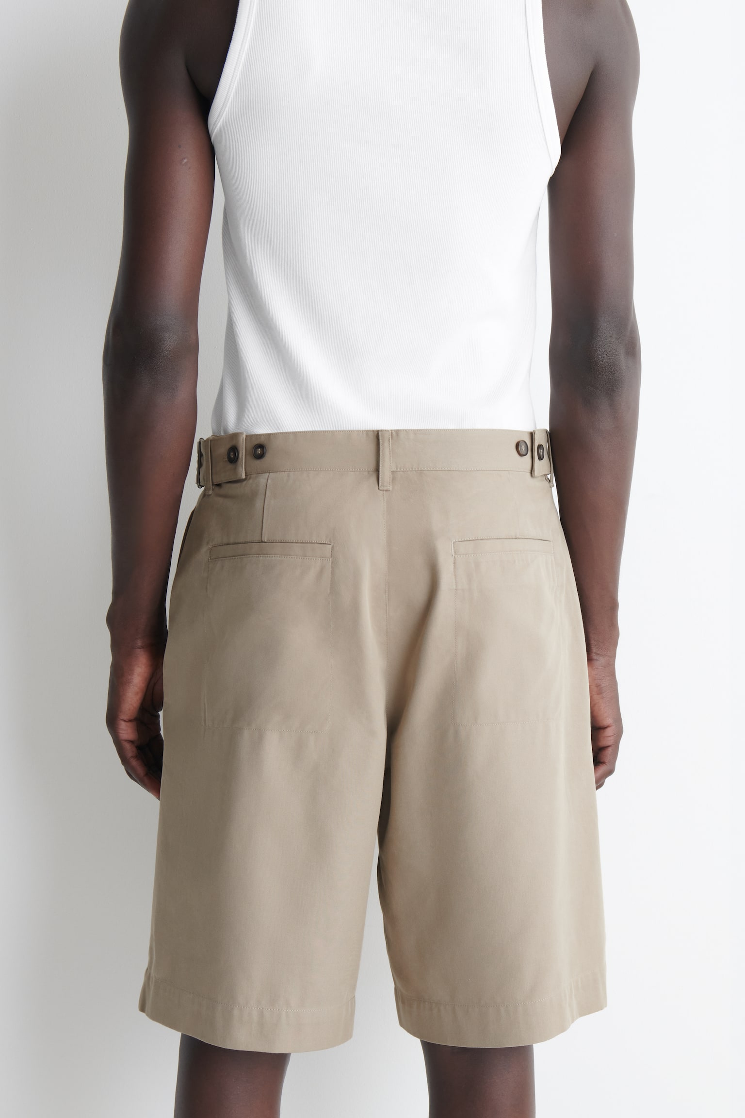 I pantaloncini beige sono mostrati in vista frontale, caratterizzati da doppie pieghe frontali e una chiusura con bottone in vita. Regolatori laterali con fibbie metalliche rettangolari dettagliano la cintura, e sono visibili tasche a fessura laterali. - 1