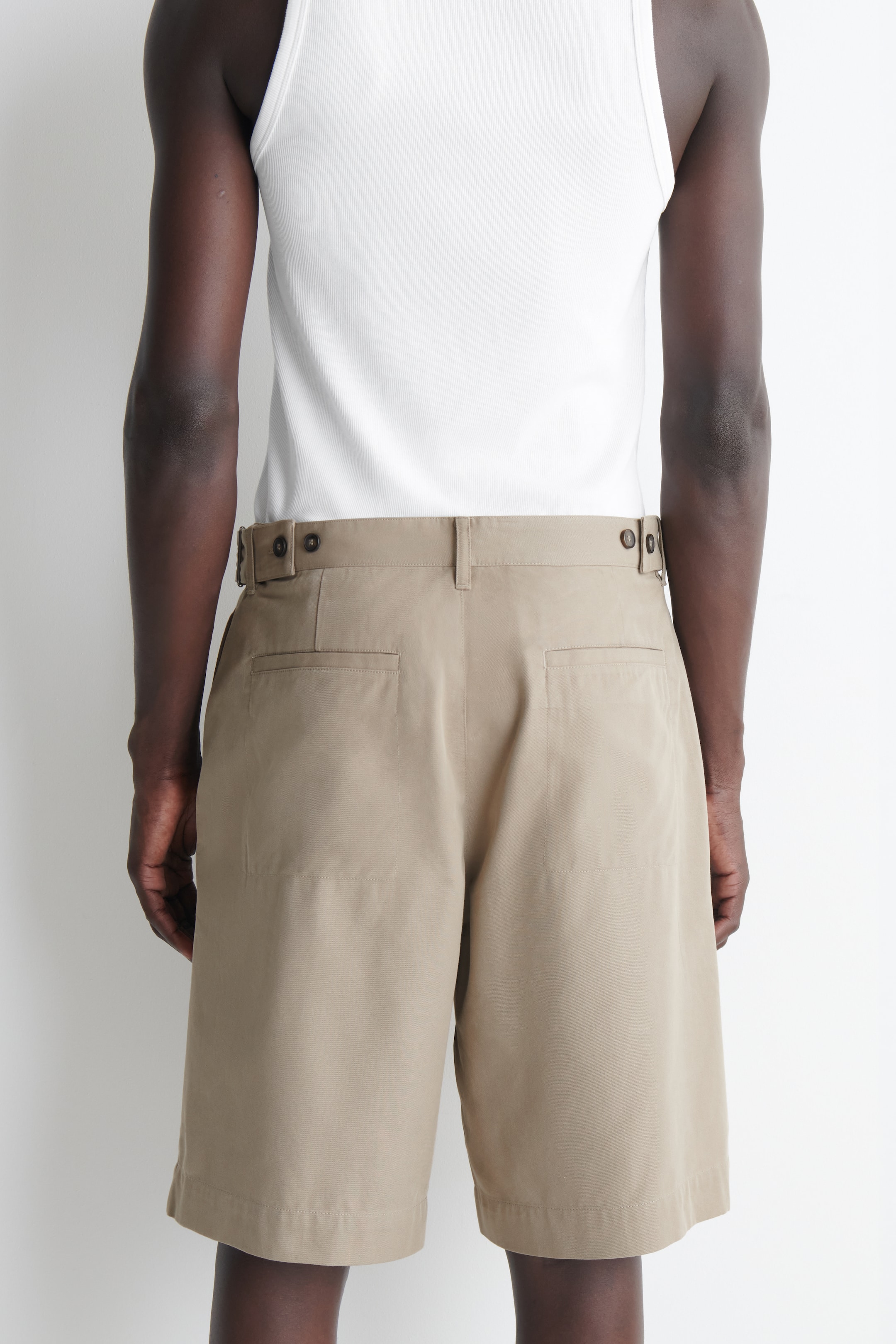 Grotere afbeelding bekijken: Kakishorts worden van de achterkant gepresenteerd en hebben een ontspannen pasvorm. Ze zijn ontworpen met knoopbare zijtabs aan de taille, twee platte achterzakken en een rechte zoom die boven de knie eindigt.