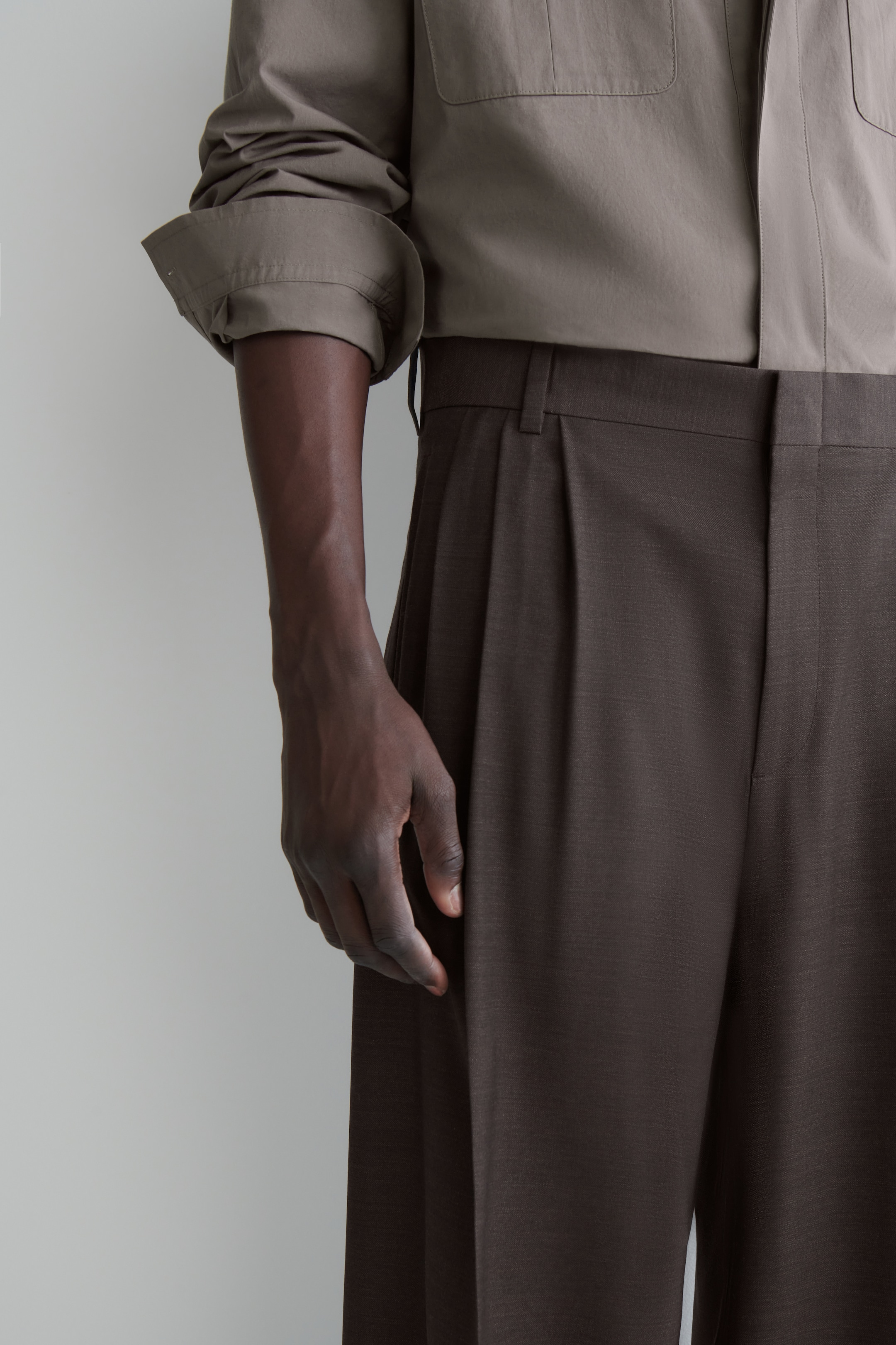 Grösseres Bild ansehen: Dunkelbraune Faltenhosen werden getragen, mit einer einzelnen Frontfalte und sichtbaren Gürtelschlaufen an der Taille. Ein hellolivfarbenes langärmliges Hemd mit aufgerolltem Ärmelbündchen wird in die Hose gesteckt.