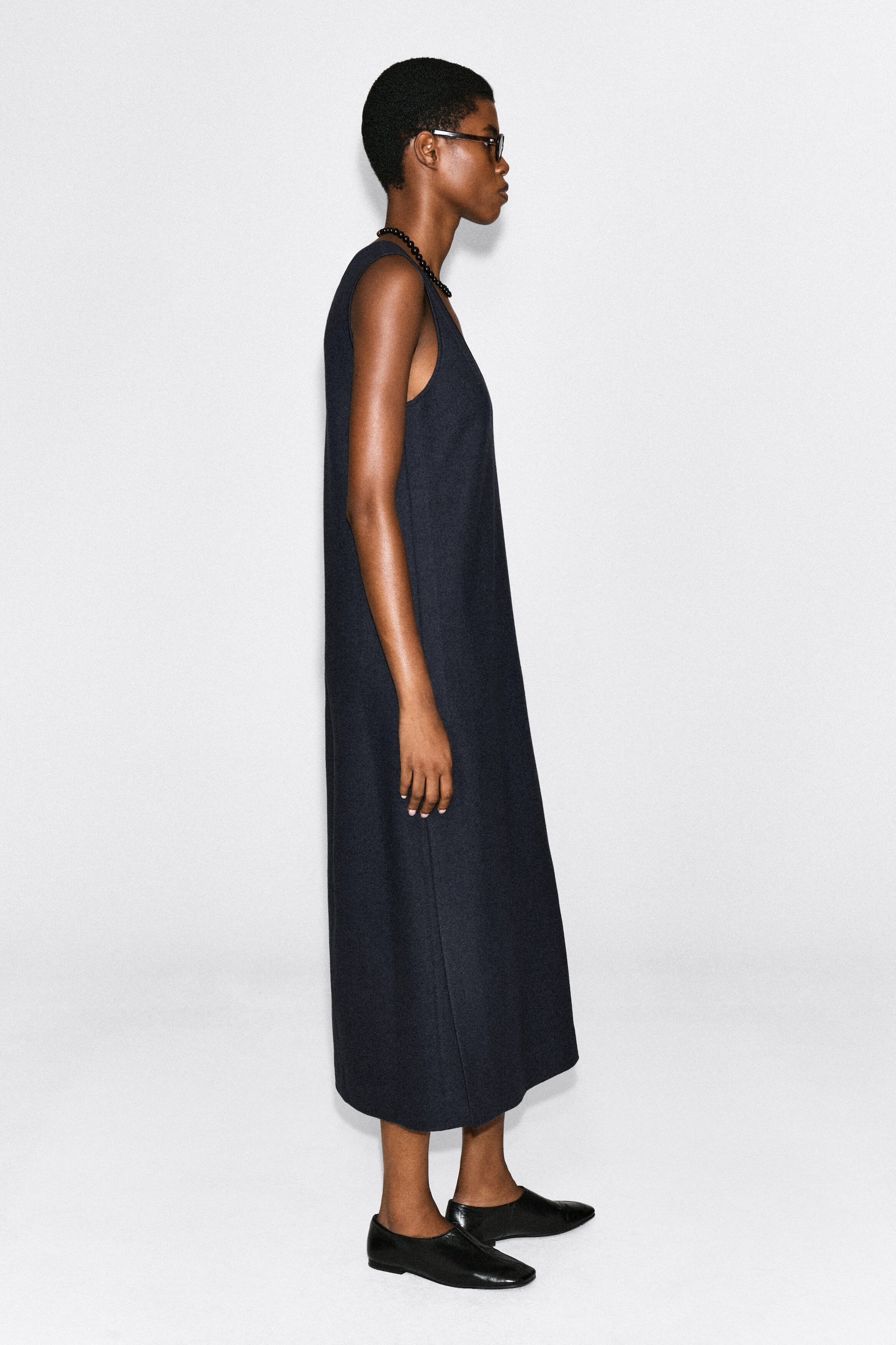 The boiled wool-blend V-neck midi dress in dark navy is worn by the model, who is standing in profile. The sleeveless, structured dress features a straight silhouette and is styled with black flat leather shoes and a beaded necklace. The boiled wool-blend V-neck midi dress in dark navy is worn by the model, who is standing in profile. The sleeveless, structured dress features a straight silhouette and is styled with black flat leather shoes and a beaded necklace.