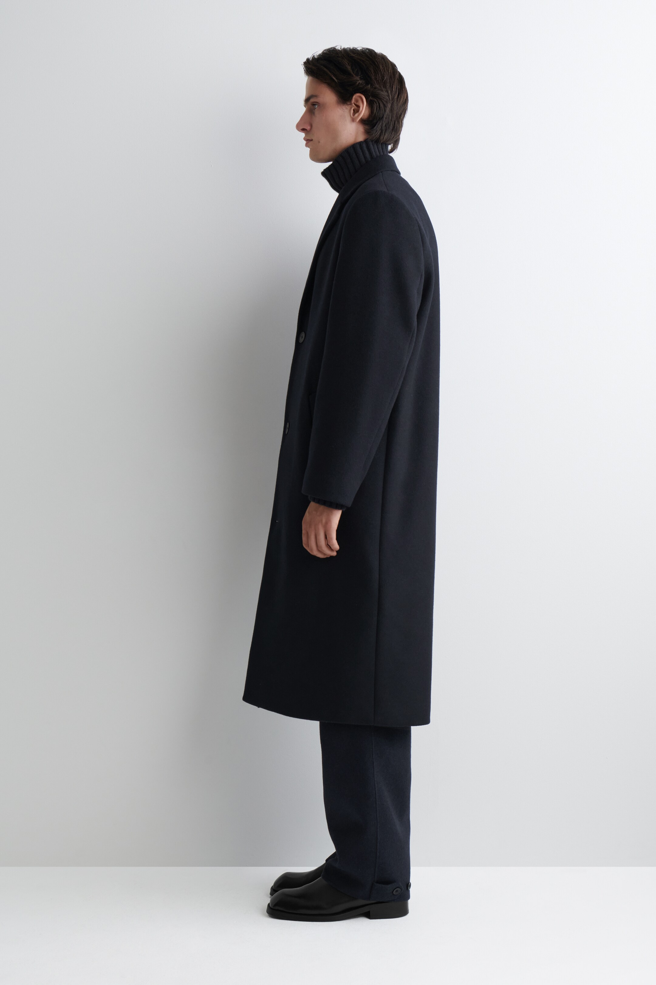View larger image: A dark overcoat is being worn in profile, facing left, featuring a smooth texture and a button-front closure. The long overcoat is extending below the knee and is layered over a dark ribbed turtleneck.