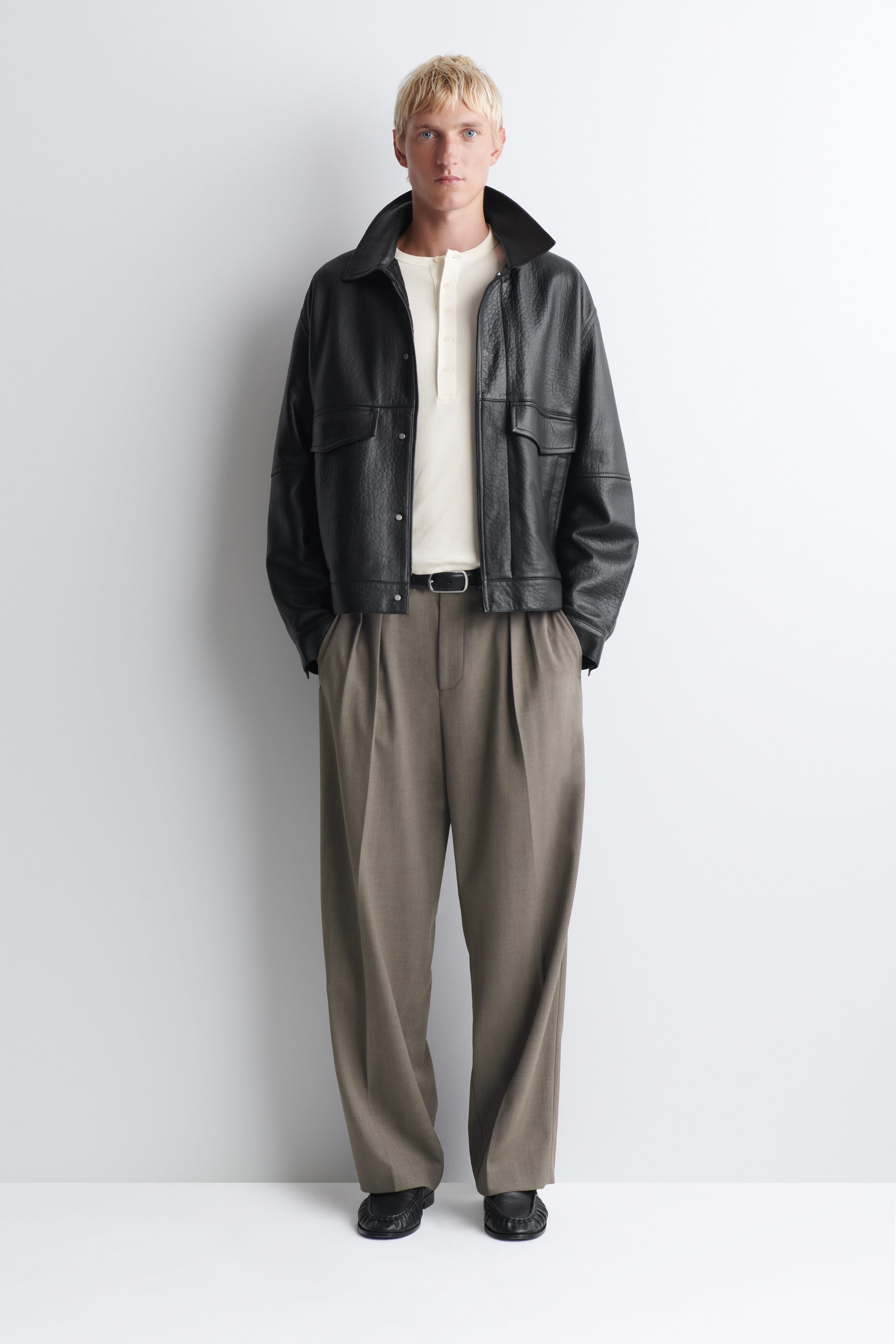 The slim-fit, cream-coloured ribbed cotton long-sleeved Henley t-shirt is shown from the front, featuring a buttoned placket at the neckline. It is styled tucked into pleated trousers and layered under an open black leather jacket. The slim-fit, cream-coloured ribbed cotton long-sleeved Henley t-shirt is shown from the front, featuring a buttoned placket at the neckline. It is styled tucked into pleated trousers and layered under an open black leather jacket.