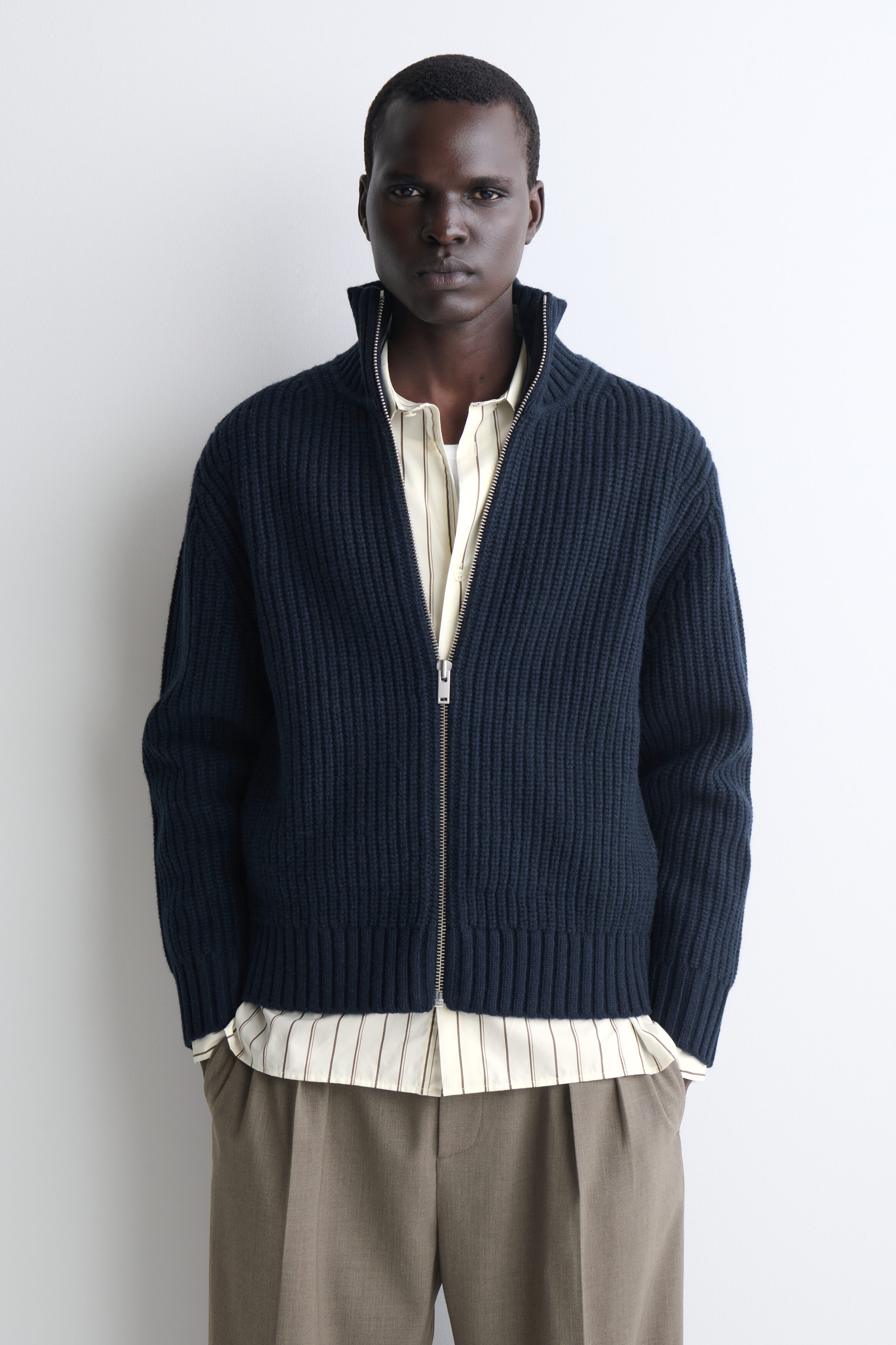 A dark navy blue ribbed-knit zip-up cardigan is shown from the front, featuring a high stand collar, a silver-tone zipper, and ribbed cuffs and hem. The cardigan is layered over a striped button-down shirt that extends past the hem, styled with pleated taupe trousers. A dark navy blue ribbed-knit zip-up cardigan is shown from the front, featuring a high stand collar, a silver-tone zipper, and ribbed cuffs and hem. The cardigan is layered over a striped button-down shirt that extends past the hem, styled with pleated taupe trousers.