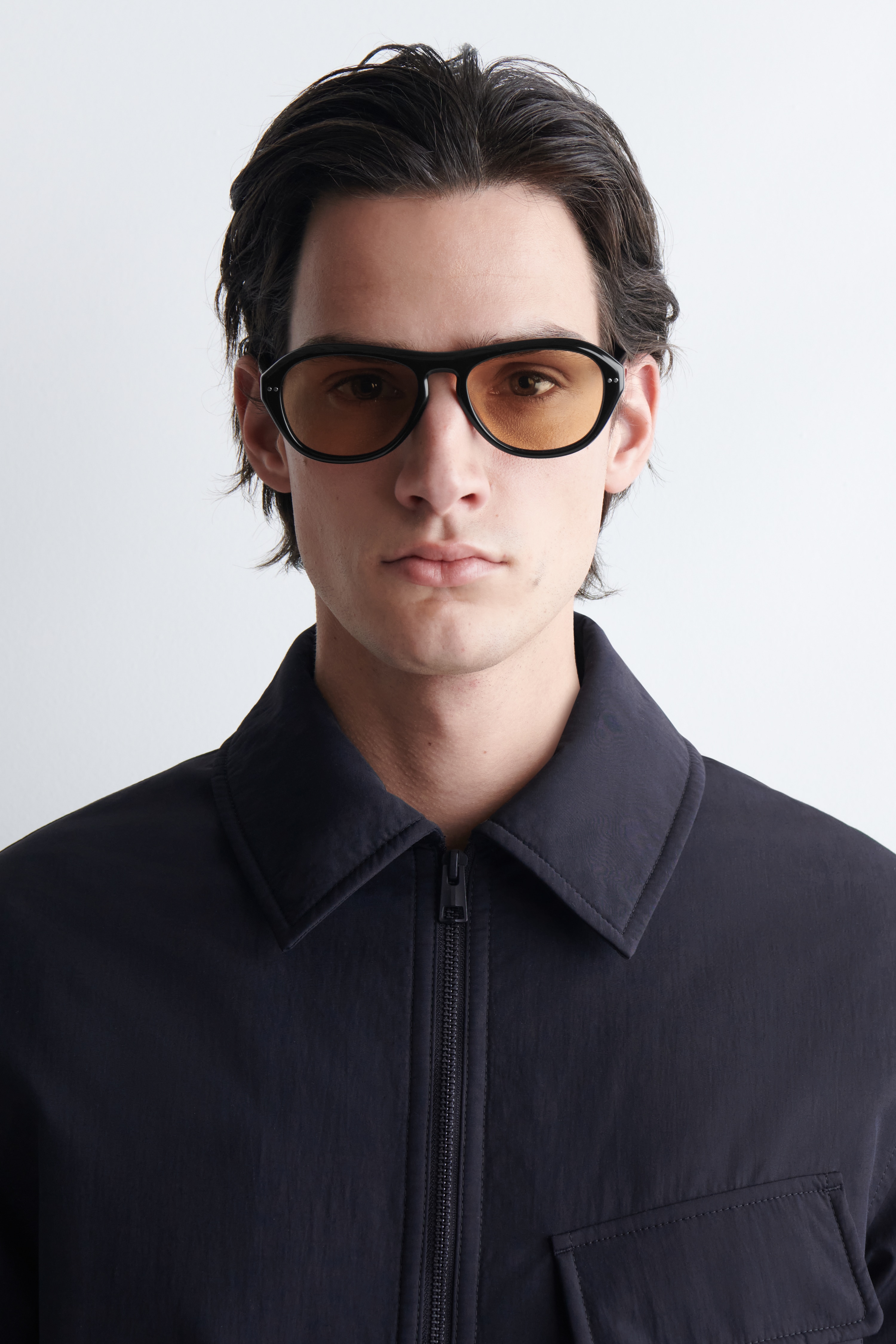 The black sunglasses are showcasing FRONT, featuring orange-tinted lenses and a slightly upswept frame with small silver accents at the upper outer corners.