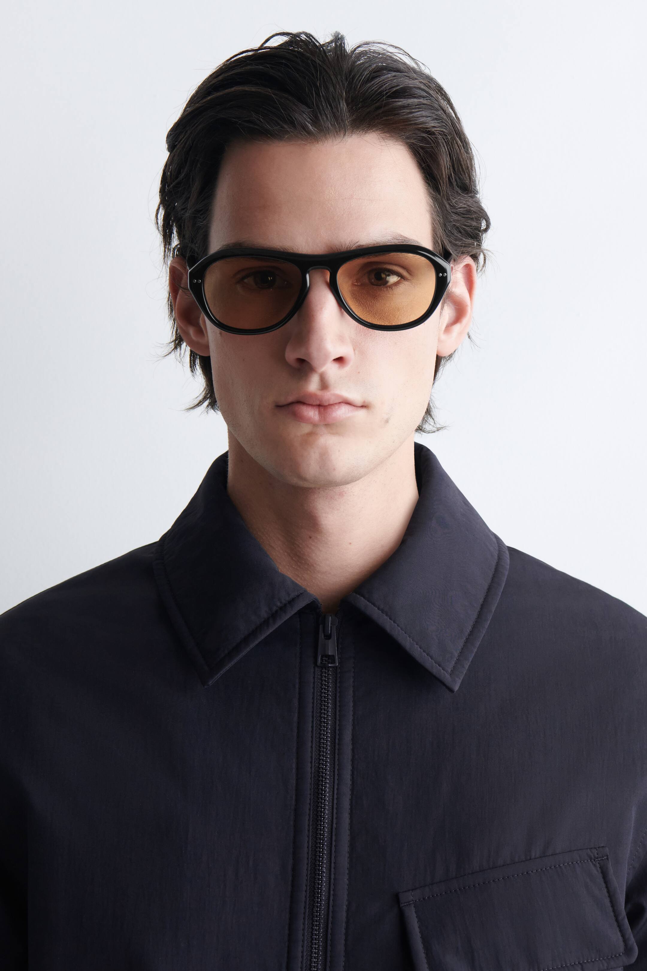 Agrandir l'image: Lunettes de soleil noires avec une monture épaisse et arrondie, des verres teintés ambre et deux petits rivets de couleur argentée à chaque coin extérieur de la monture avant. Elles sont portées face à l'avant.
