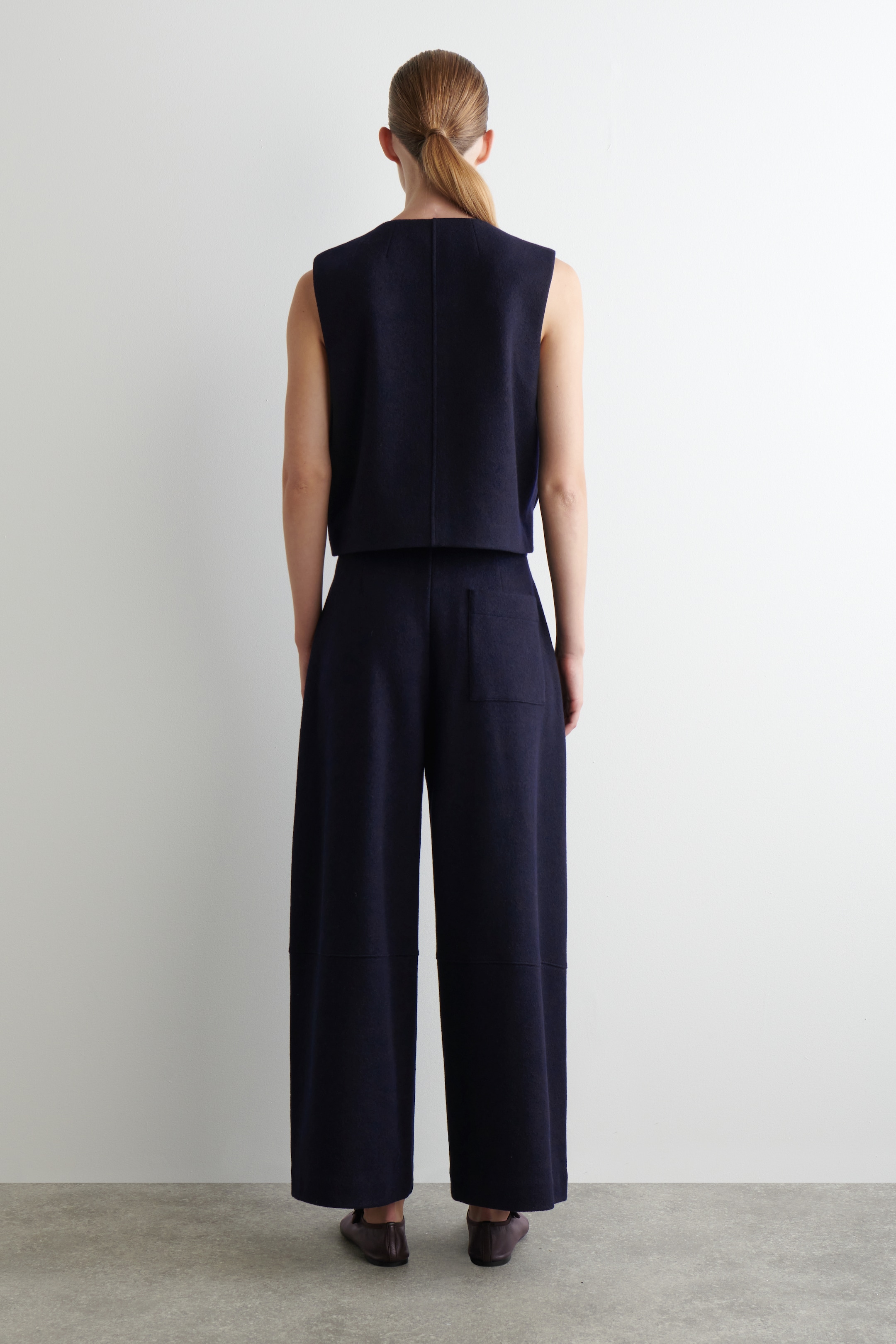 Ingrandisci l'immagine: Un completo coordinato blu navy a due pezzi viene presentato di spalle su una modella. Il top senza maniche presenta una cucitura verticale al centro della schiena, e i pantaloni a gamba larga mostrano una tasca applicata sulla destra di chi li indossa e cuciture orizzontali a pannelli, con scarpe scure e piatte che completano il look.