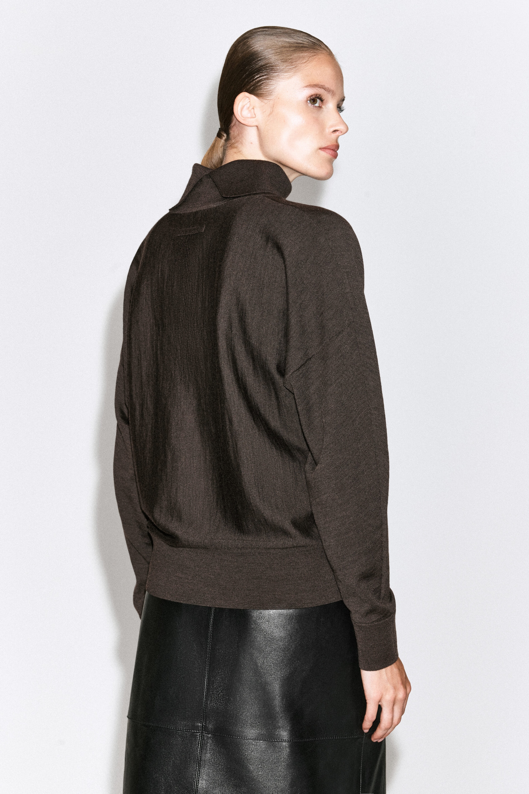 A dark brown, high-neck knitted jumper featuring long sleeves and a contrasting sheer panel down the centre back is being worn by the model. The jumper is styled with a black leather skirt. A dark brown, high-neck knitted jumper featuring long sleeves and a contrasting sheer panel down the centre back is being worn by the model. The jumper is styled with a black leather skirt.