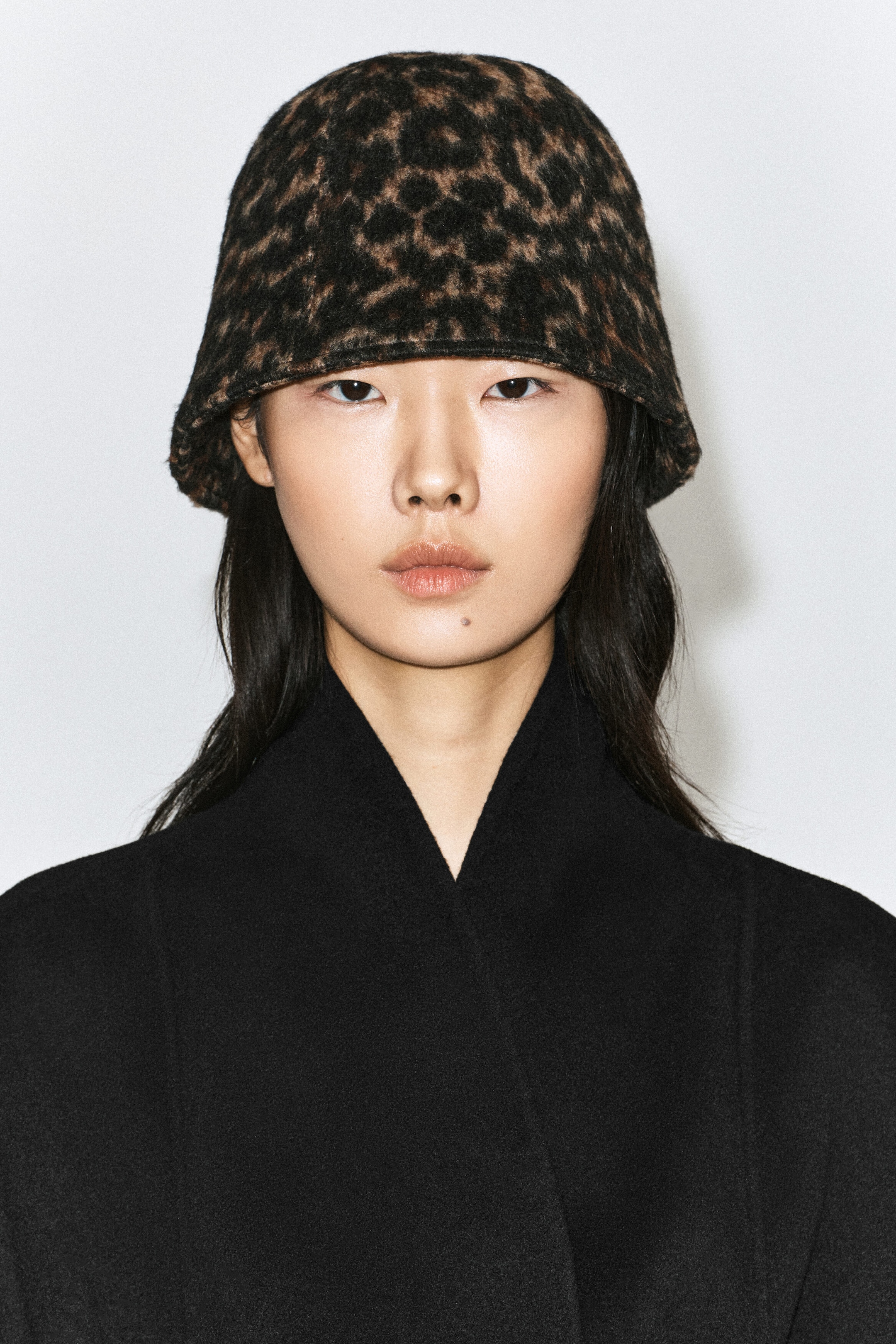 A leopard-print boiled wool bucket hat with a textured finish and a gently curved brim is being worn by the model. The hat is styled with a black high-collared coat. A leopard-print boiled wool bucket hat with a textured finish and a gently curved brim is being worn by the model. The hat is styled with a black high-collared coat.