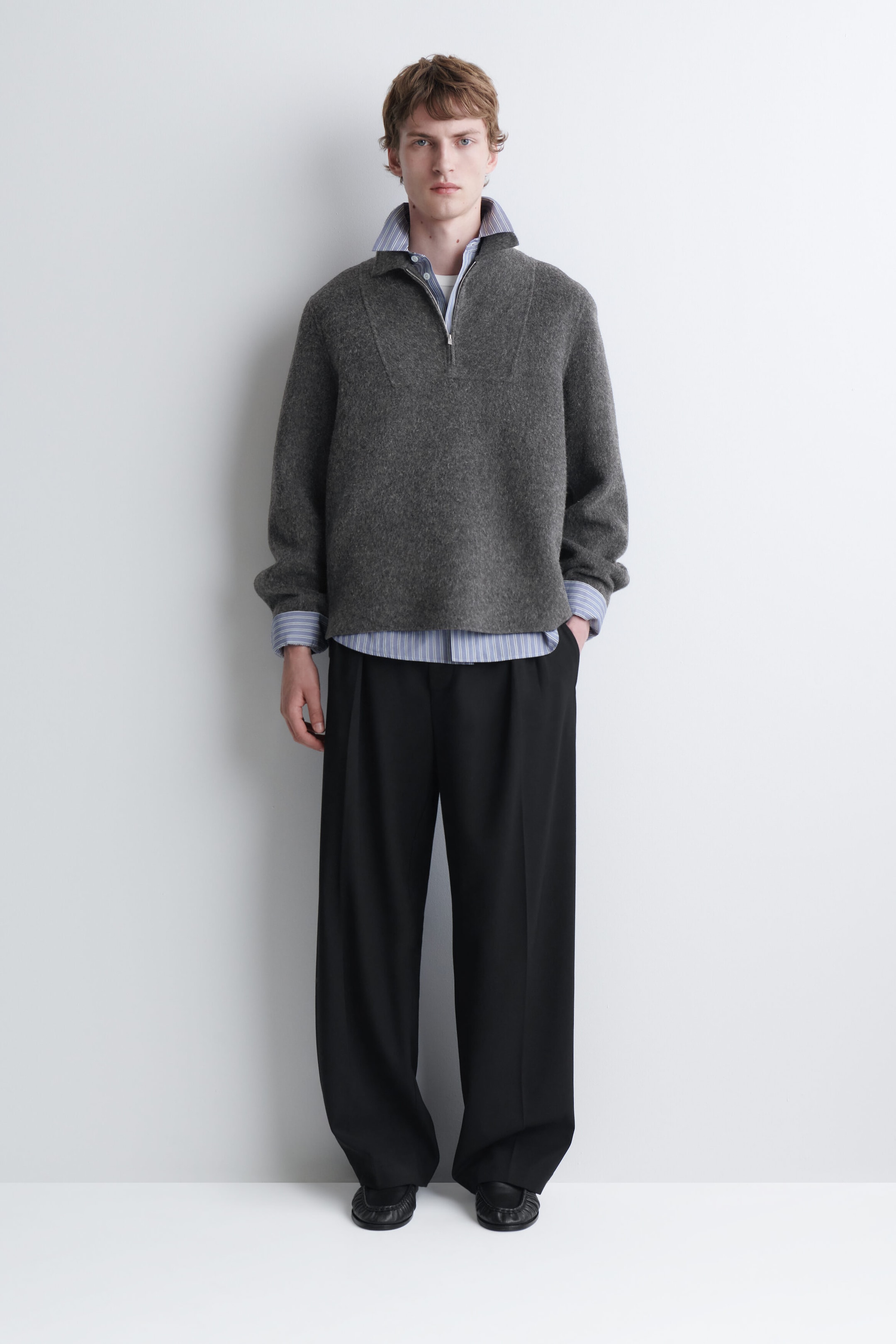 A gray, textured knit half-zip sweater is being worn by the model, layered over a blue-and-white striped button-down shirt. The sweater features a high collar with a partial front zipper and is styled with wide-leg black trousers.