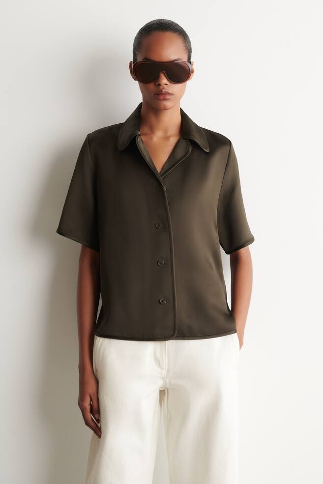 Una camisa verde oscuro se presenta de frente, con mangas cortas, cuello tipo campamento y cierre completo de botones al frente con botones oscuros a juego. La camisa exhibe un tejido suave y lustroso y un ajuste relajado. - 1