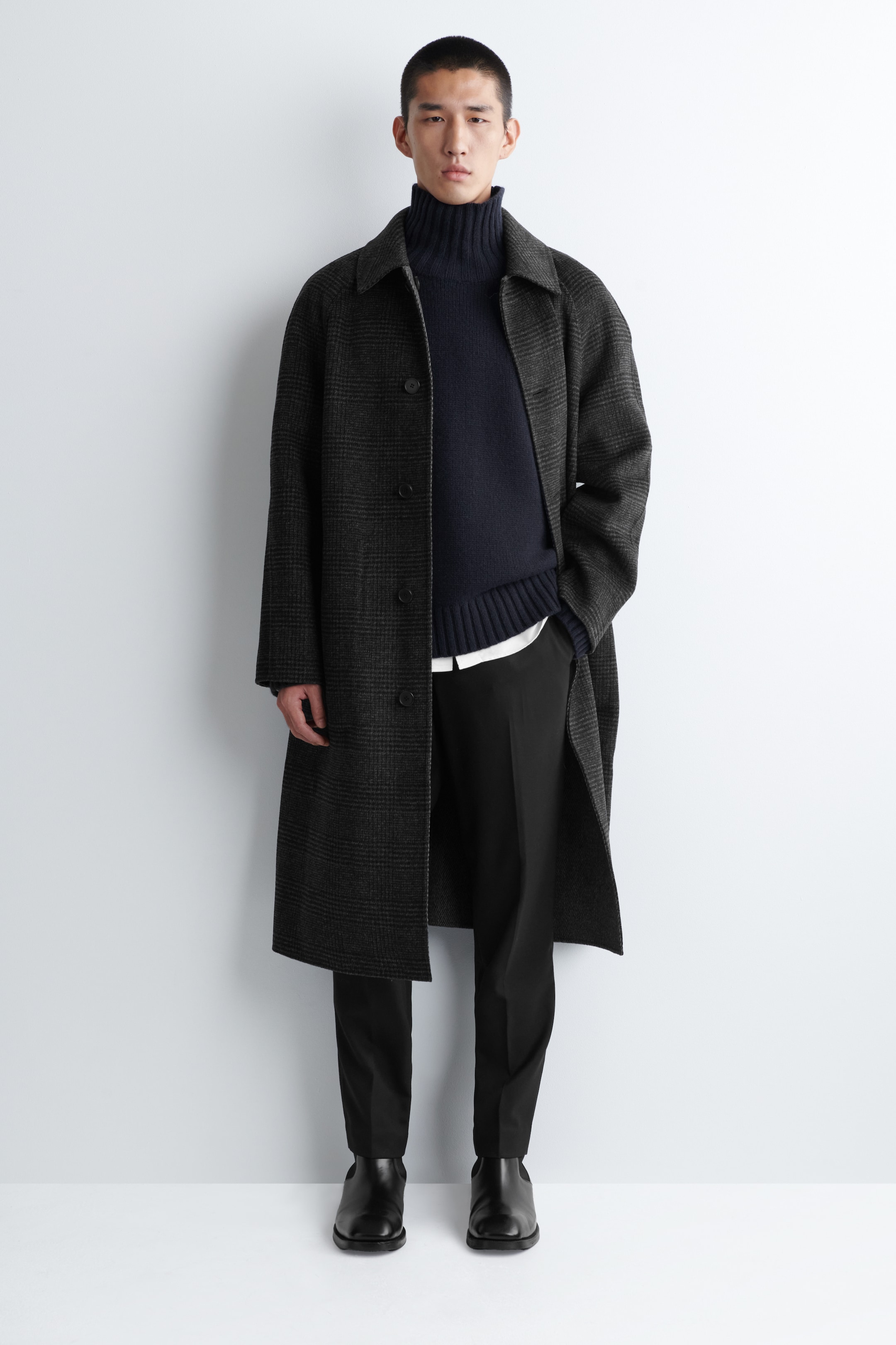 Visualizza immagine più grande: Un abito da uomo è presentato in una vista frontale, caratterizzato da un lungo cappotto a quadri grigio scuro con colletto a punta, indossato aperto sopra un maglione dolcevita a costine blu navy e una camicia con colletto bianco, completato da pantaloni neri e stivaletti in pelle nera.