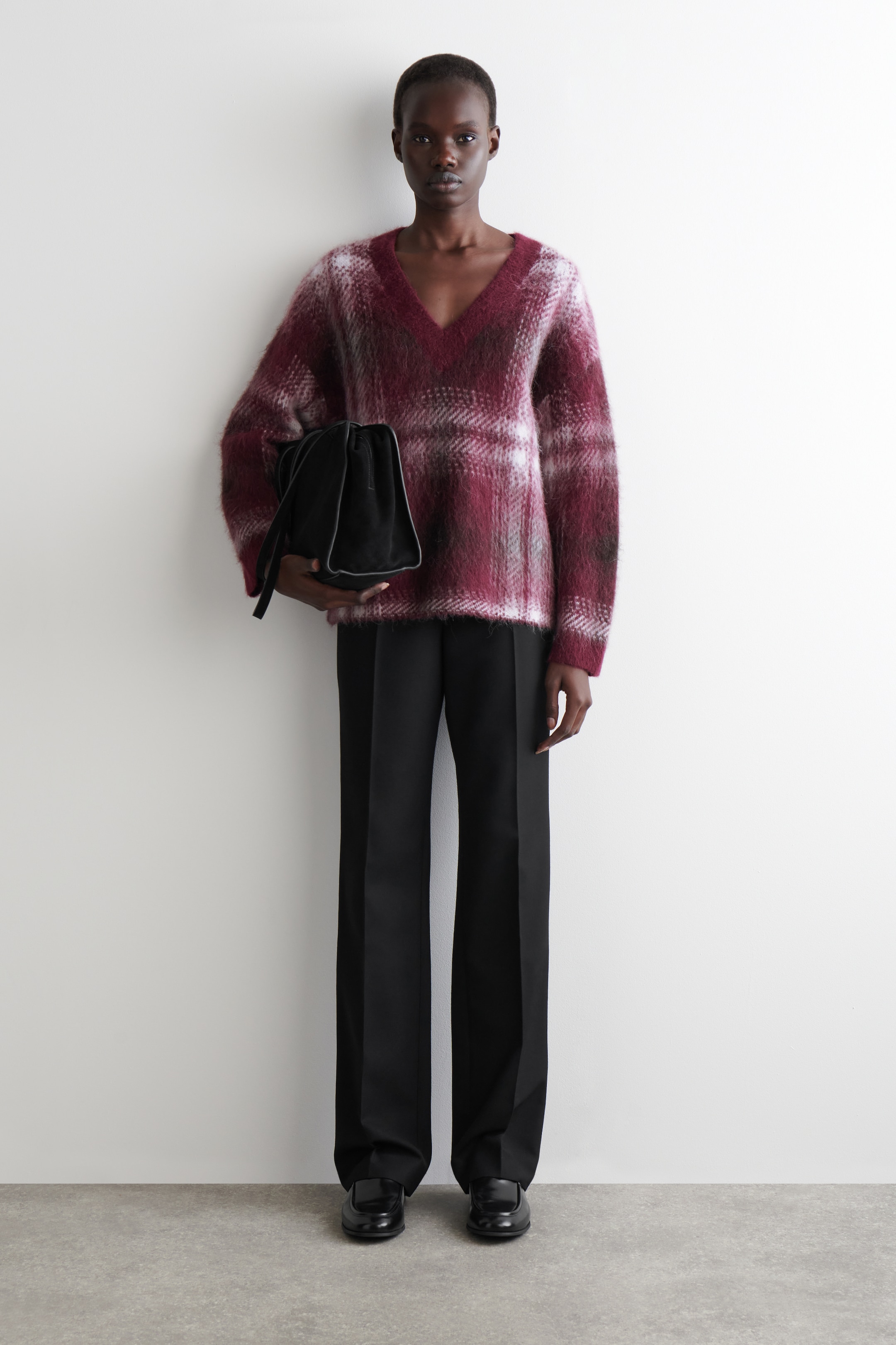 Grösseres Bild ansehen: Eine Ganzkörperansicht eines Outfits mit einem flauschigen V-Ausschnitt-Pullover, der ein Karomuster in Burgunderrot, Weiß und Dunkelgrau zeigt. Dies wird mit schwarzen, weit geschnittenen Hosen und schwarzen Loafer-Schuhen gestylt. Eine schwarze, weiche Beuteltasche wird in der Hand gehalten.
