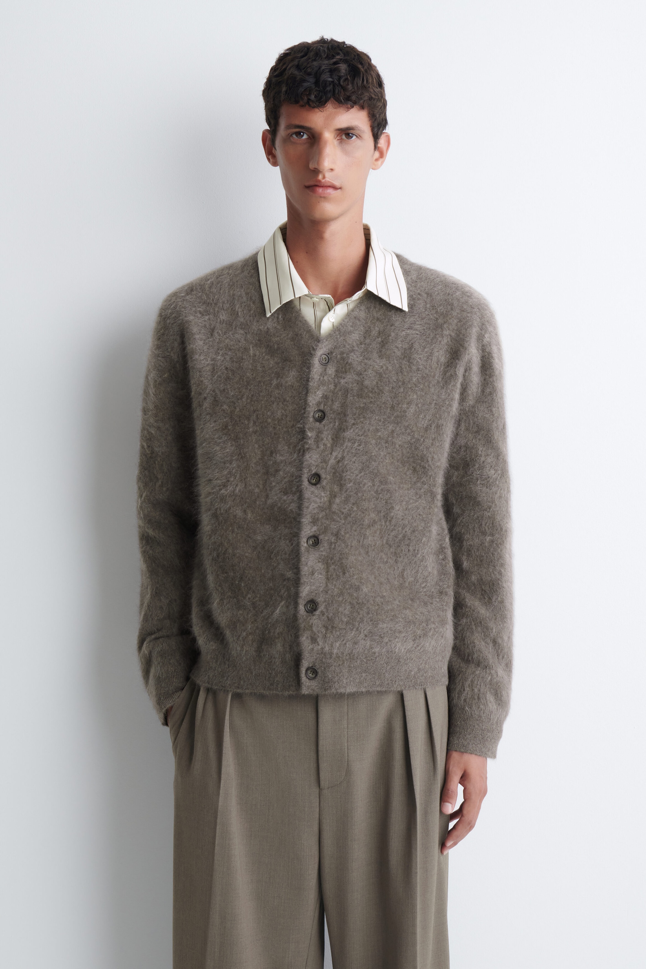 Un cardigan à col en V en cachemire brossé, tricot duveteux gris taupe, est porté par le mannequin par-dessus une chemise rayée à col. Le cardigan est entièrement boutonné sur le devant et agencé avec un pantalon plissé brun olive. Un cardigan à col en V en cachemire brossé, tricot duveteux gris taupe, est porté par le mannequin par-dessus une chemise rayée à col. Le cardigan est entièrement boutonné sur le devant et agencé avec un pantalon plissé brun olive.