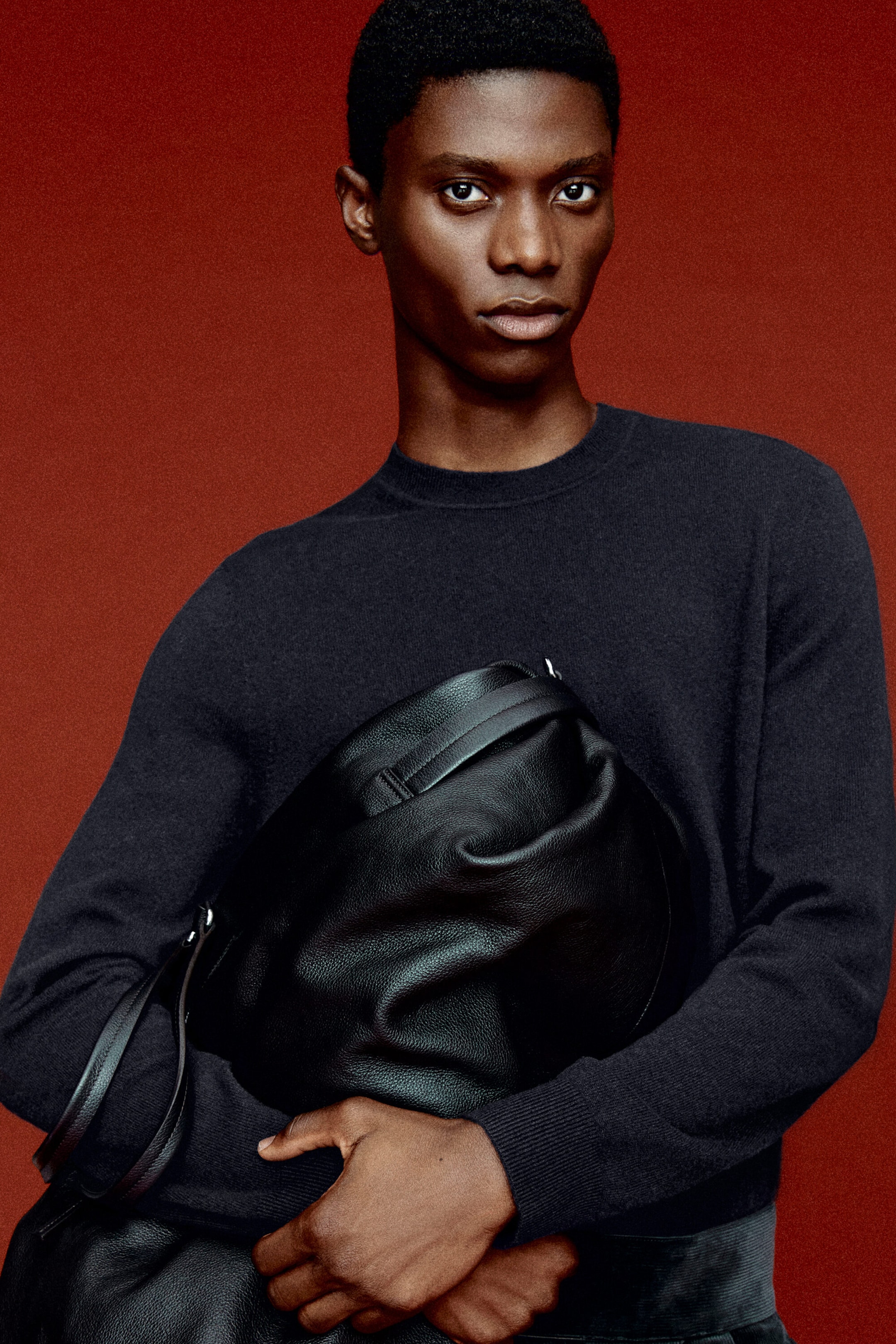 Un mannequin de face porte un chandail en cachemire bleu marine foncé, doté d’un col rond côtelé et de poignets côtelés, tout en tenant un sac de voyage en cuir grainé noir. Le sac en cuir souple, porté de façon décontractée, est tenu devant, mettant en valeur son grain texturé et sa bandoulière en cuir.