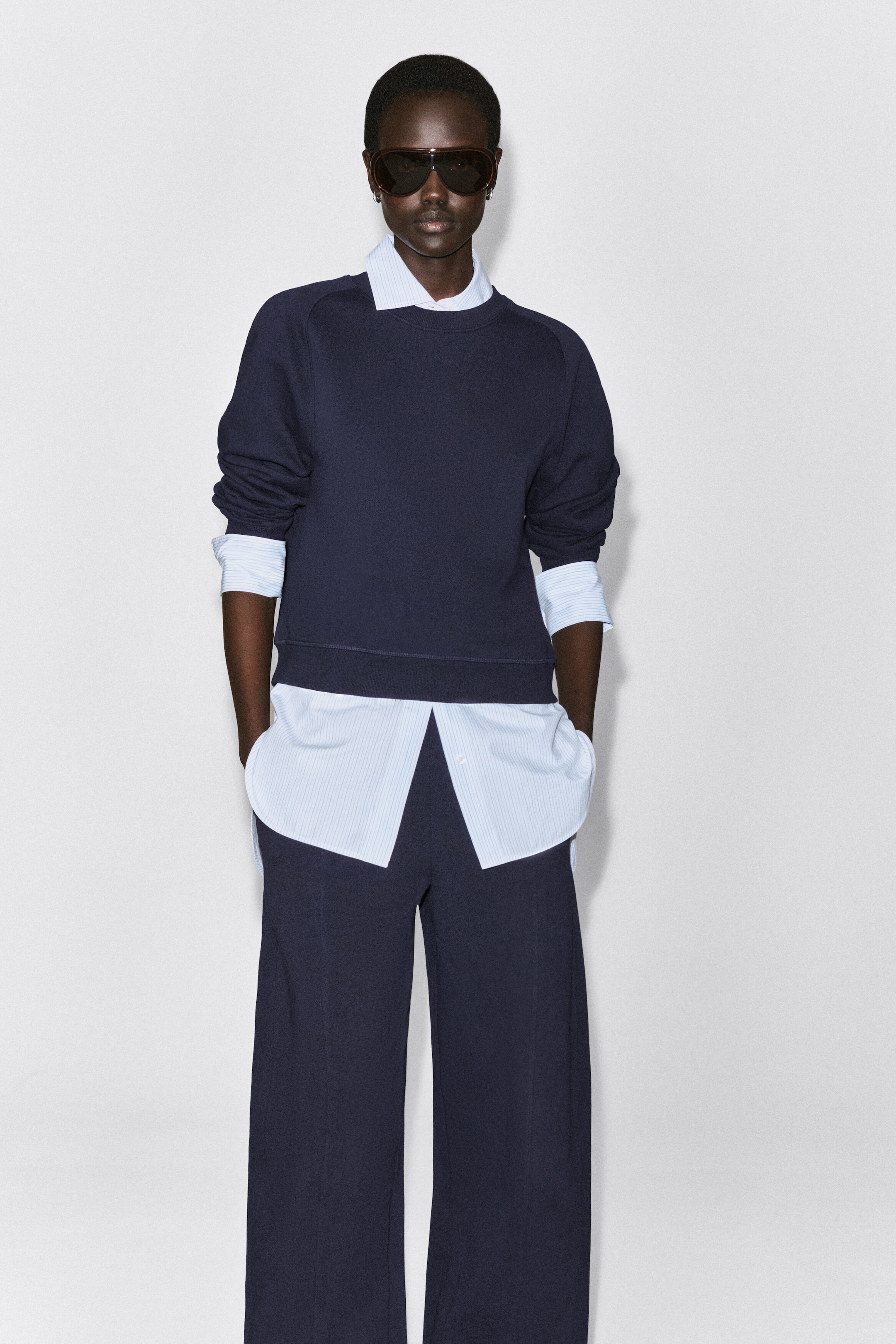 A navy blue cotton sweatshirt is being shown from the front, featuring a relaxed fit, a ribbed crewneck, raglan sleeves, and a ribbed hem. The sweatshirt is layered over a light blue striped collared shirt with visible cuffs and hem, and is paired with matching navy trousers. A navy blue cotton sweatshirt is being shown from the front, featuring a relaxed fit, a ribbed crewneck, raglan sleeves, and a ribbed hem. The sweatshirt is layered over a light blue striped collared shirt with visible cuffs and hem, and is paired with matching navy trousers.