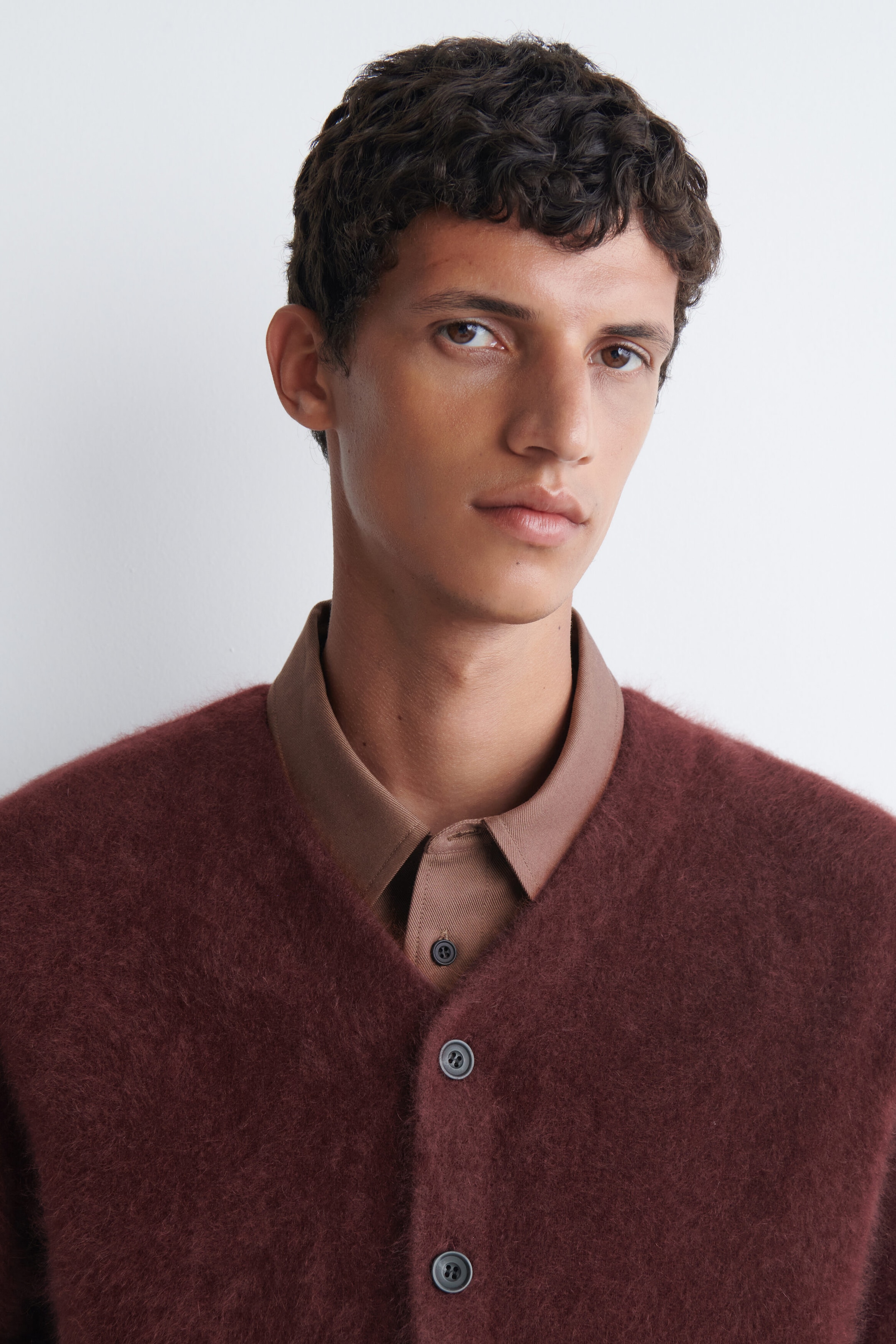 A burgundy brushed-cashmere V-neck cardigan is being worn by the model over a brown collared shirt. The cardigan features a soft, textured finish and dark front buttons. A burgundy brushed-cashmere V-neck cardigan is being worn by the model over a brown collared shirt. The cardigan features a soft, textured finish and dark front buttons.