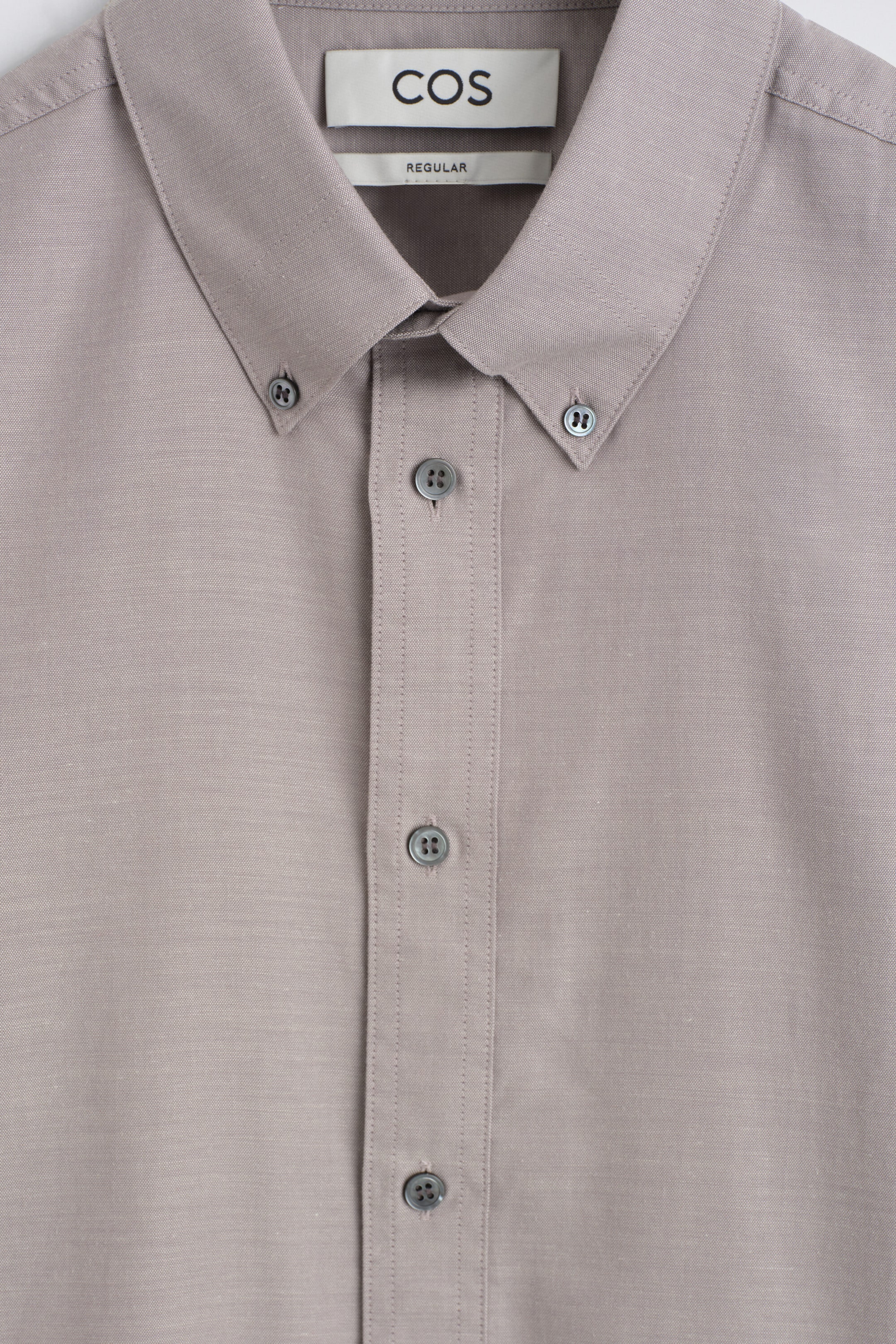 A close-up front view of the button-down collar brushed Oxford shirt shows its soft taupe-grey fabric and structured collar design. The shirt features a classic button-down collar fastened with small grey buttons, a central button placket, and a visible COS brand label indicating a regular fit.