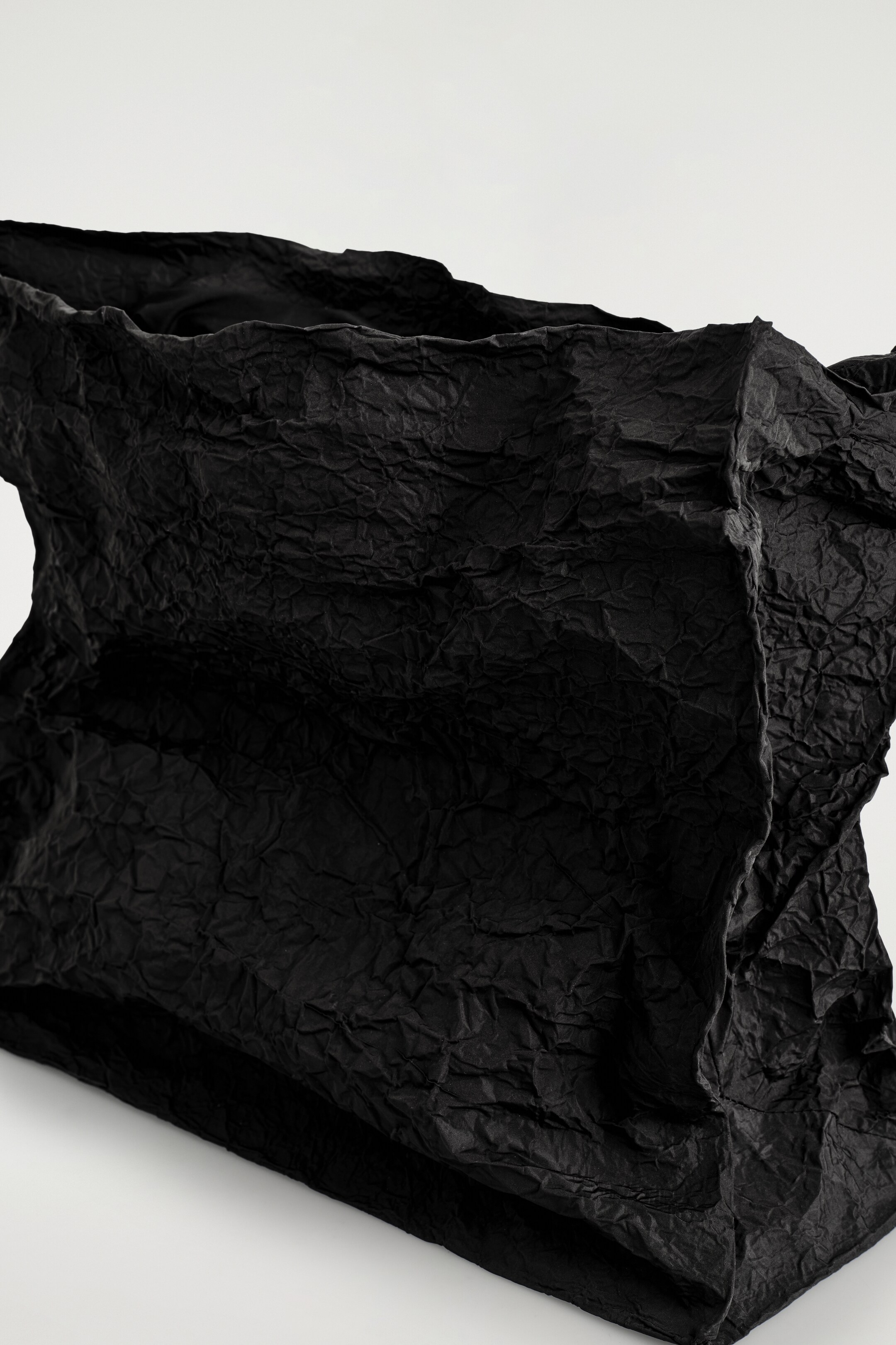 Grösseres Bild ansehen: Eine übergroße schwarze Clutch wird mit der oberen Öffnung zum Betrachter präsentiert. Sie zeichnet sich durch eine stark zerknitterte Stoffstruktur aus, die eine übertriebene, unstrukturierte Silhouette erzeugt.