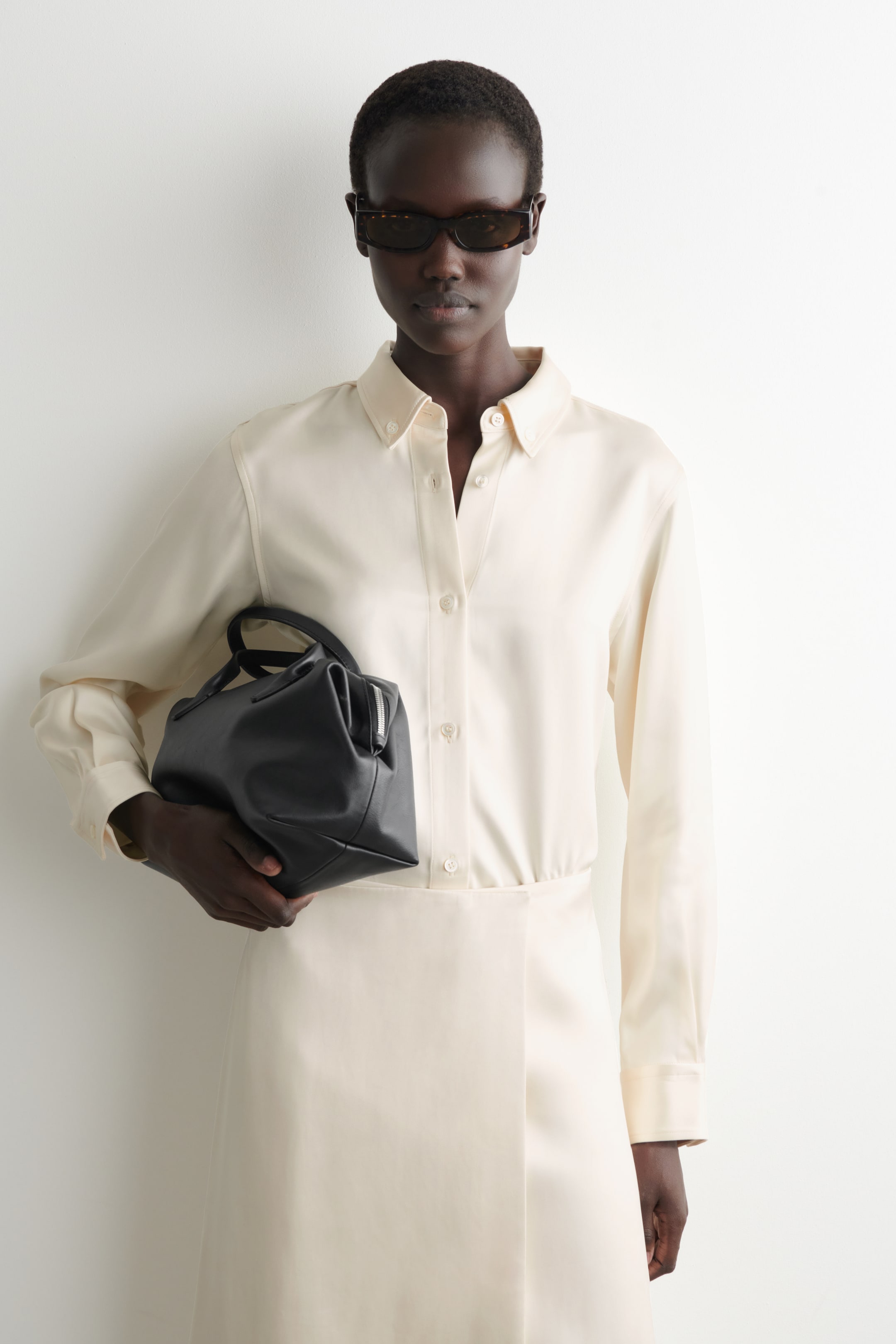 Zobraziť väčší obrázok: Čierna kabelka je držaná, pričom je viditeľná jej predná a bočná strana. Má štruktúrovaný, ale mäkký obdĺžnikový tvar, integrované horné rukoväte a strieborný zips pozdĺž horného okraja.