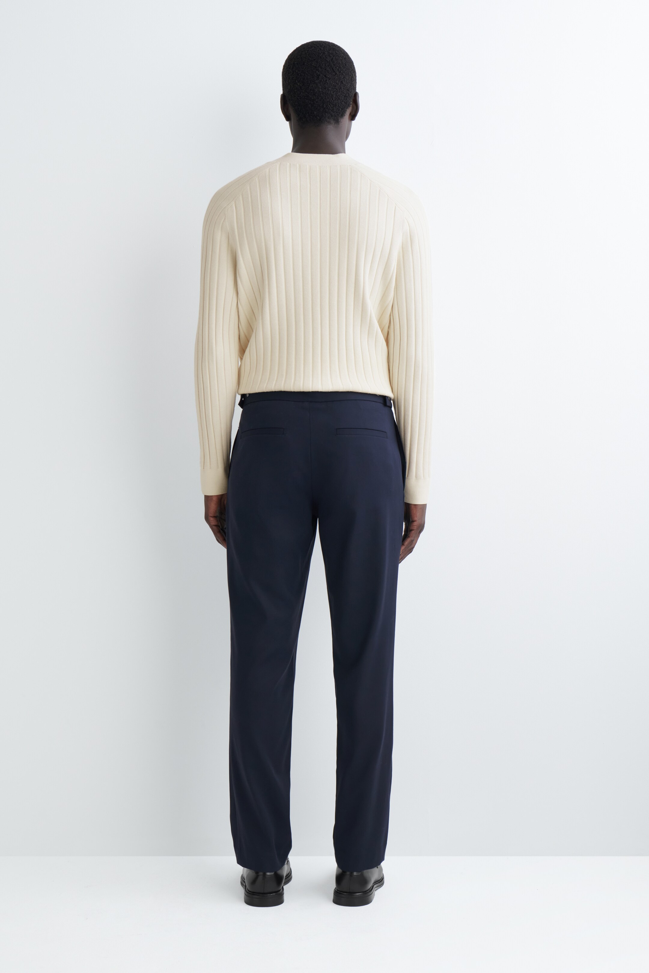View larger image: A cream ribbed knit sweater and navy trousers are being presented from a full back view. The sweater is featuring vertical ribbing and long sleeves, while the trousers are showcasing a straight-leg fit with subtle back welt pockets.