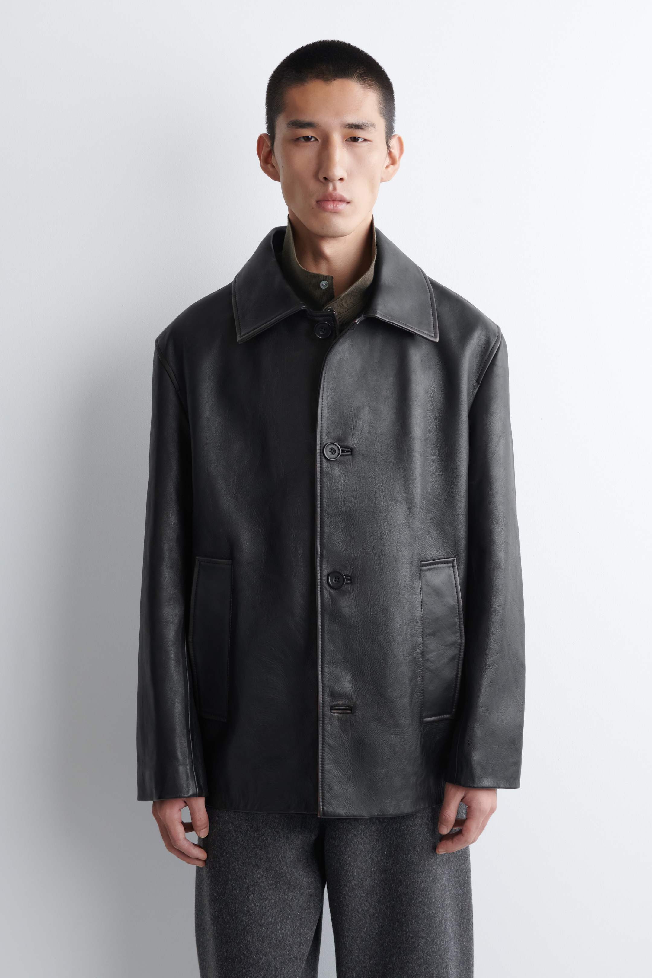 Agrandir l'image: Une veste en cuir noir est présentée de face, avec un col pointu classique sur une chemise verte visible. La veste est conçue avec trois boutons sombres sur le devant, deux grandes poches plaquées, et des coutures plus claires contrastantes le long de ses coutures et bords, affichant une coupe décontractée.
