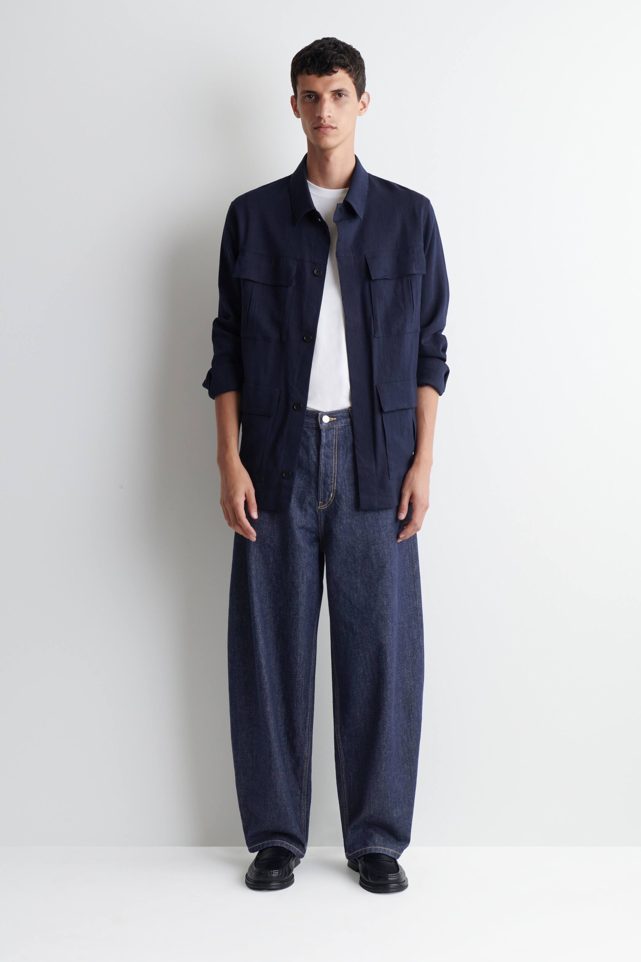 Grotere afbeelding bekijken: De donker indigo barrel-leg Mallow jeans worden gedragen door het model. Ze hebben een ontspannen, gebogen pasvorm en zichtbare tabakskleurige stiksels. De jeans worden gestyled met een wit t-shirt en een donkerblauw overhemdjack.