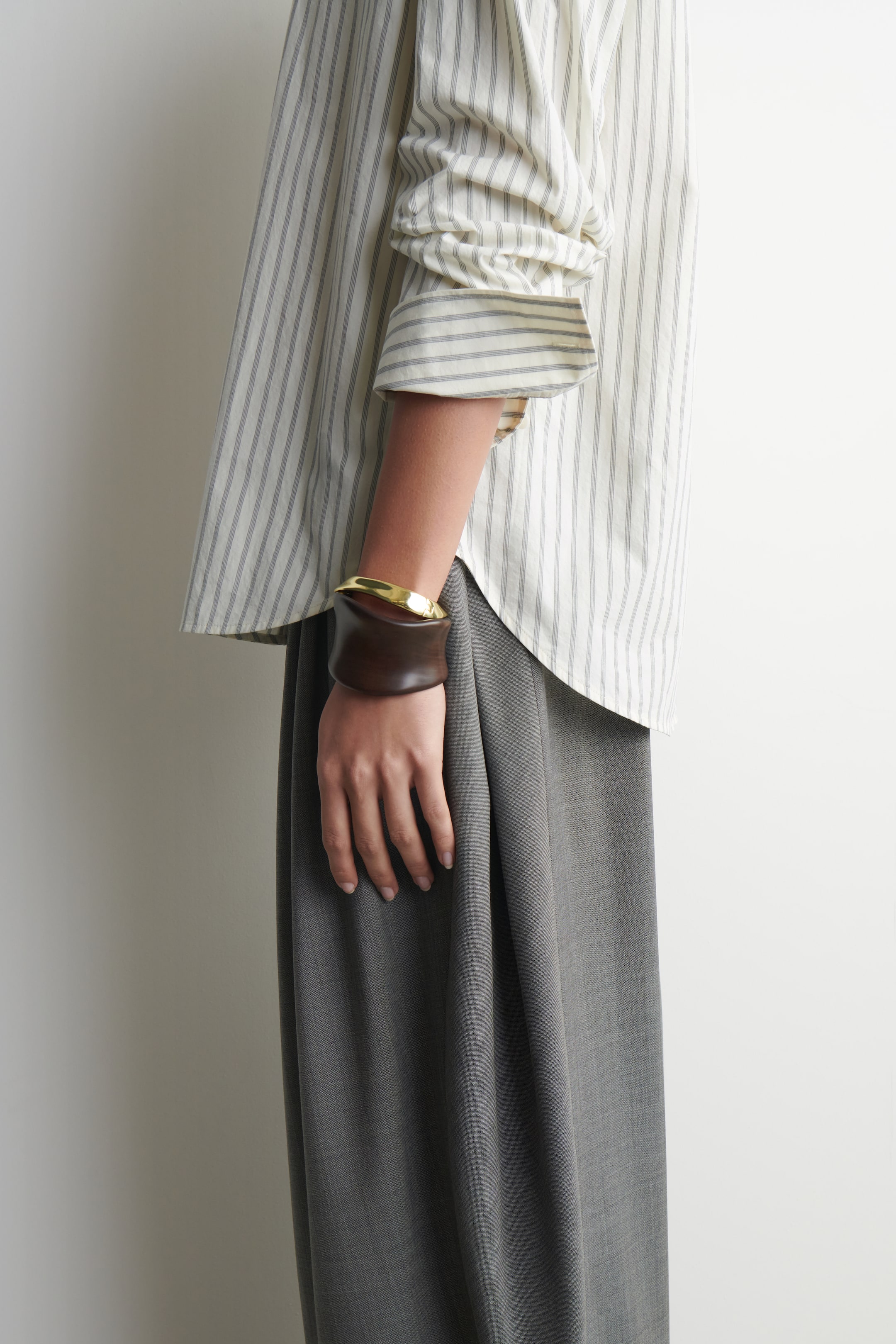 Visualizza l'immagine ingrandita: Una camicia color crema con sottili righe verticali grigie presenta una vestibilità rilassata con maniche lunghe e polsini abbottonati, che scivola su pantaloni grigi a gamba larga. Un ampio bracciale scultoreo, che combina una sezione curva scura con un accento metallico dorato lucido, viene indossato sopra il polsino della camicia sull'avambraccio sinistro.